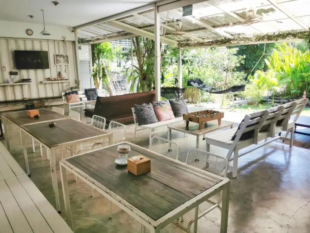 Bright airy patio with rustic wooden tables, white chairs, and cozy couches. A foosball table sits centrally. Greenery and hammock outside create a relaxed, inviting vibe.