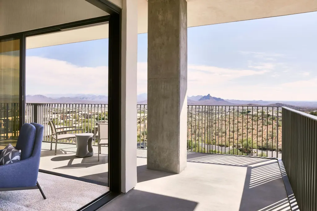 Modern balcony with sliding glass doors opening to a spacious patio. Overlooks a scenic desert landscape with distant mountains under a clear sky.