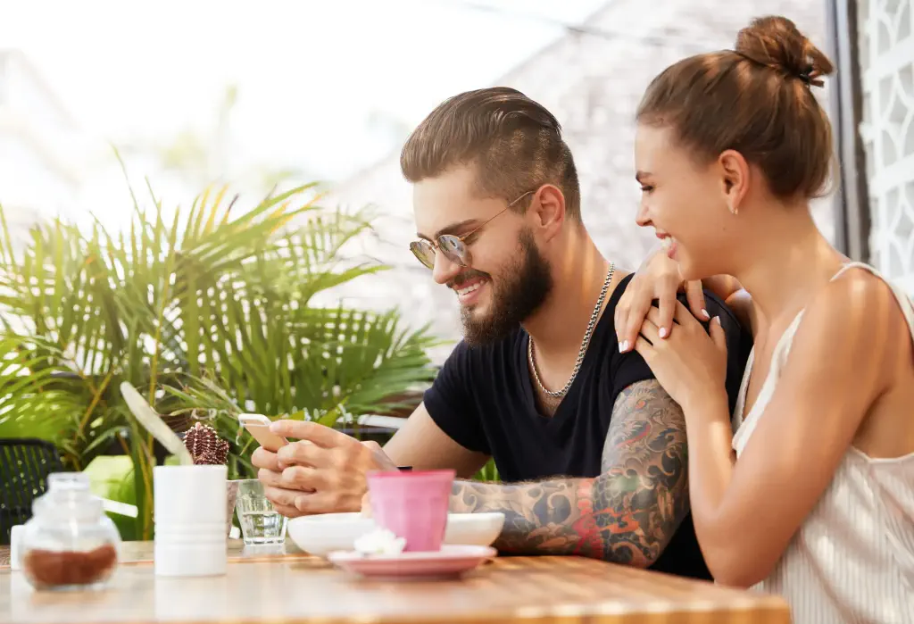 A tattooed man using his smartphone as a woman looks over his shoulder.