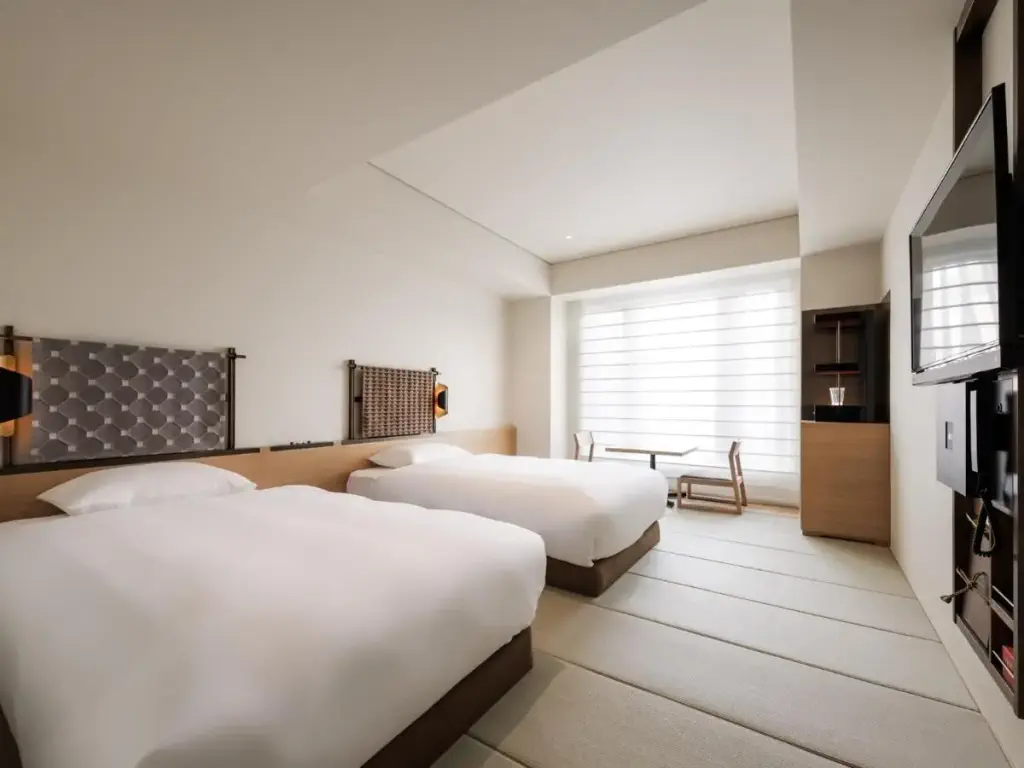 Modern hotel room with two neatly made single beds, light wood accents, a flat-screen TV, and a small table with chairs by a large window. 