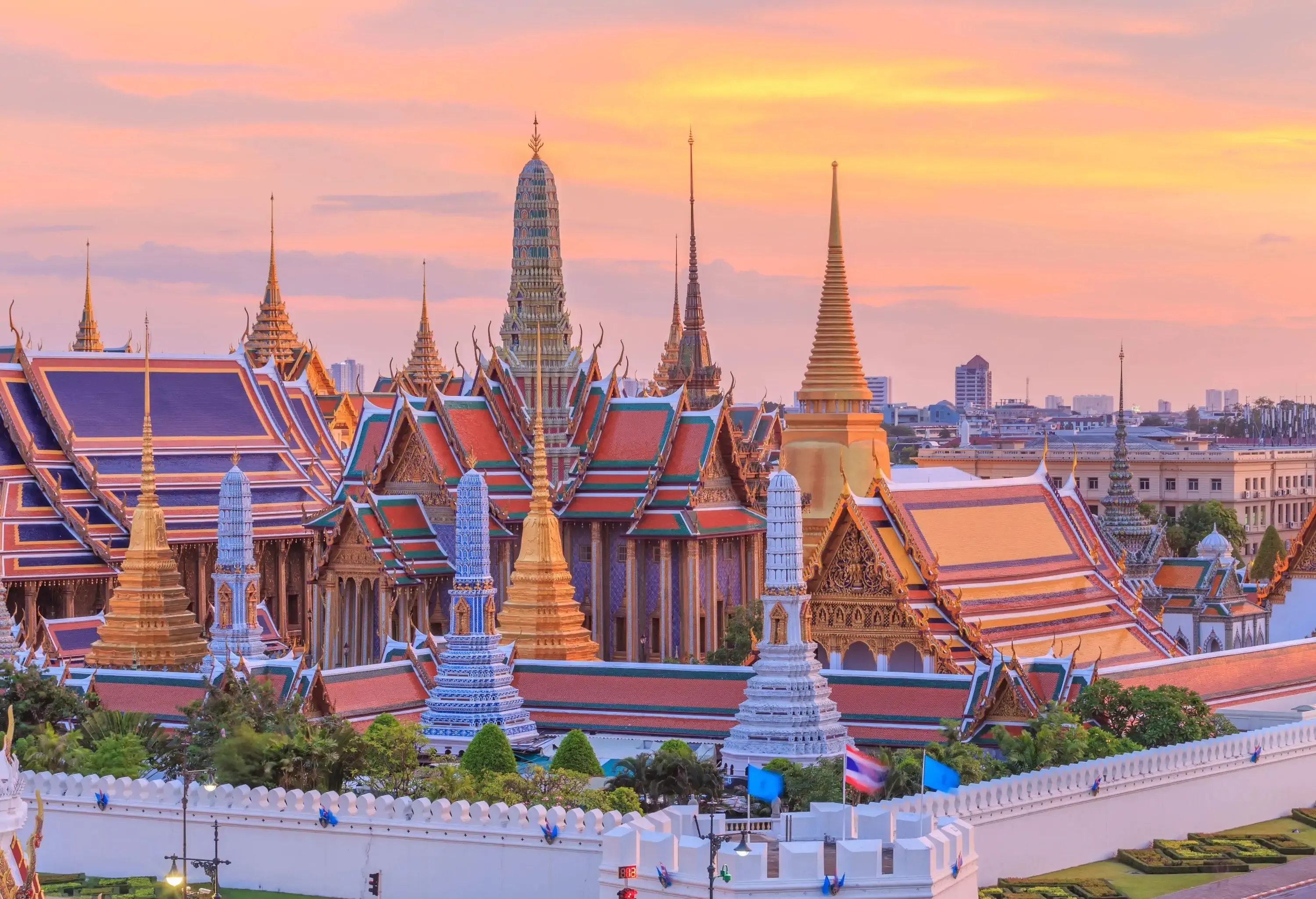 The Grand Palace's exquisitely crafted golden temples are surrounded by white ceramic tiled structures with towering spires and golden bell-shaped stupas under the setting sun.