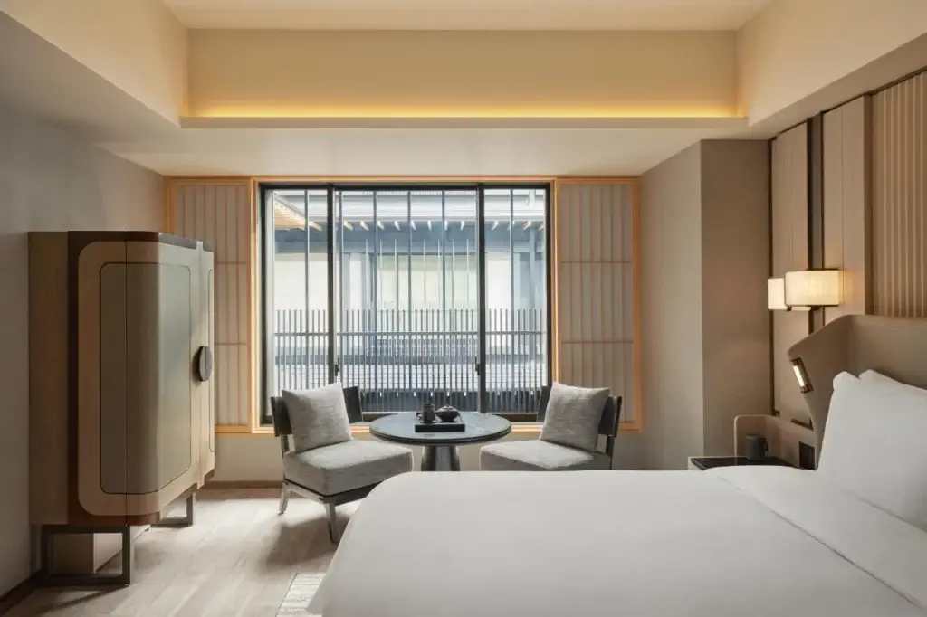 Modern hotel room with minimalistic decor. Features a large bed, two chairs, and a round table by a window with lattice detail.