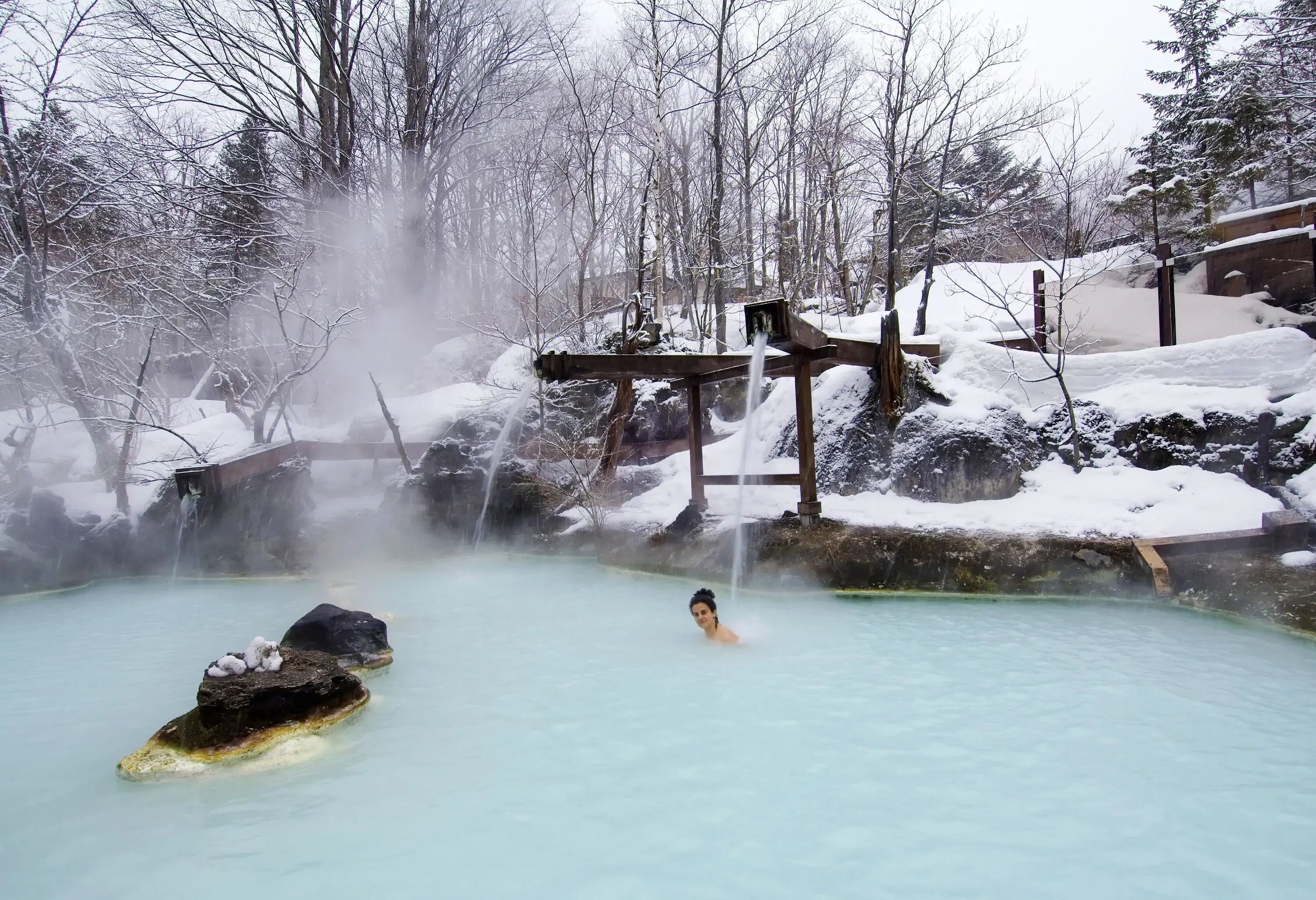 A woman dips into a steaming outdoor hot spring surrounded by frosted trees and snow-covered landscapes.