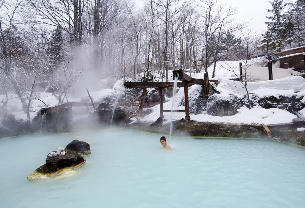 A woman dips into a steaming outdoor hot spring surrounded by frosted trees and snow-covered landscapes.