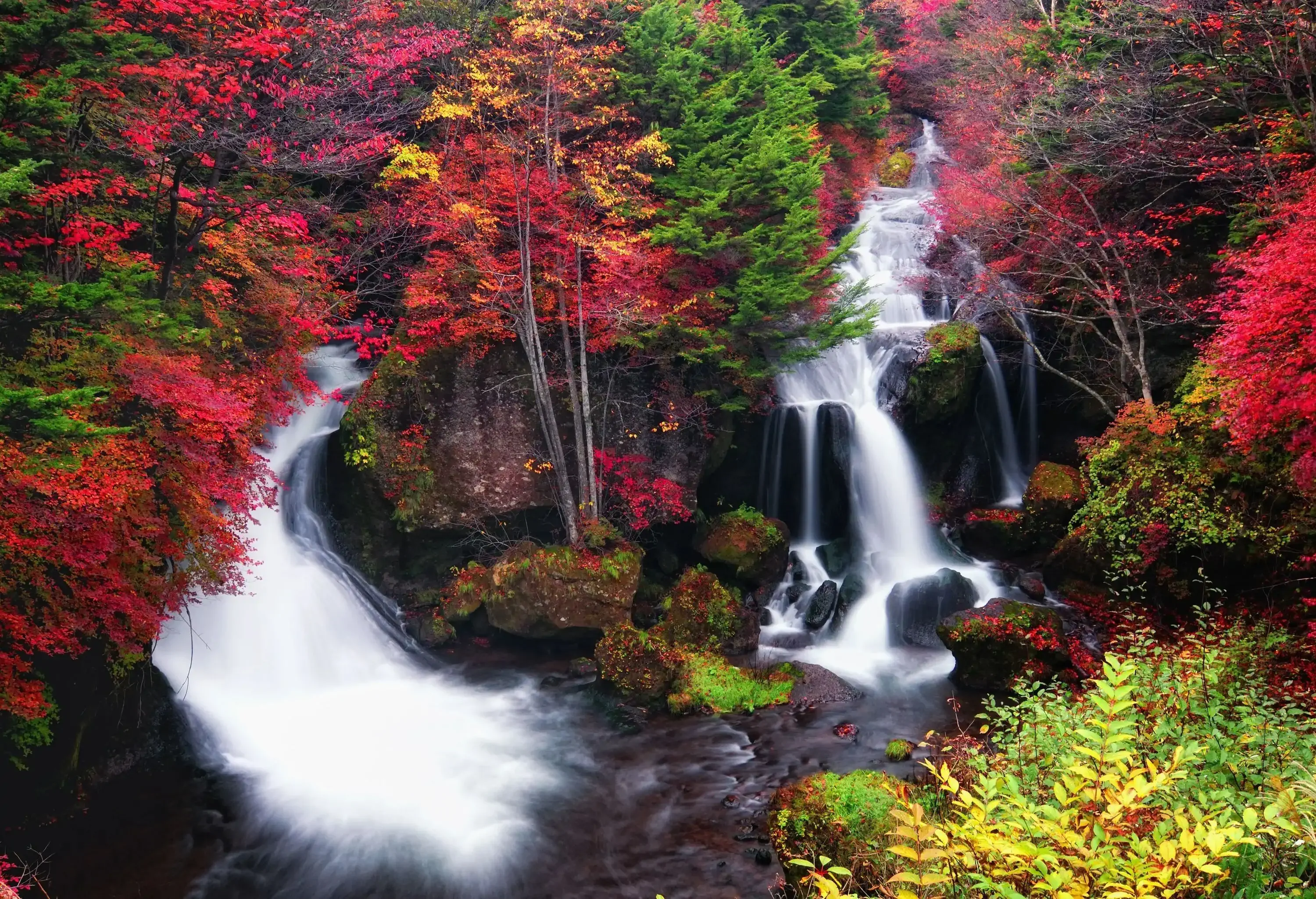 Two waterfalls cascade into a rocky stream surrounded by vibrant autumn foliage.