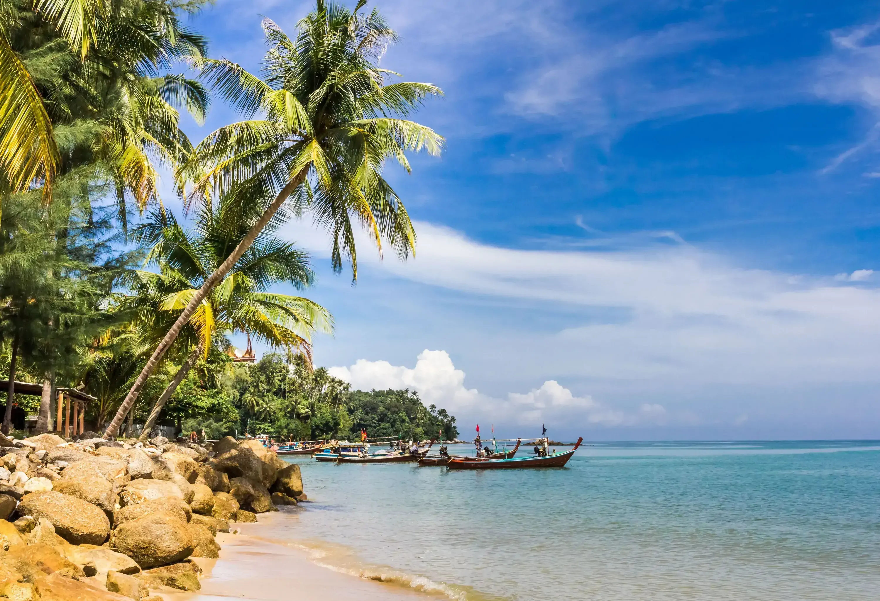 Boats moored on the tranquil waters of the beach lined with lush trees and a pile of rocks.