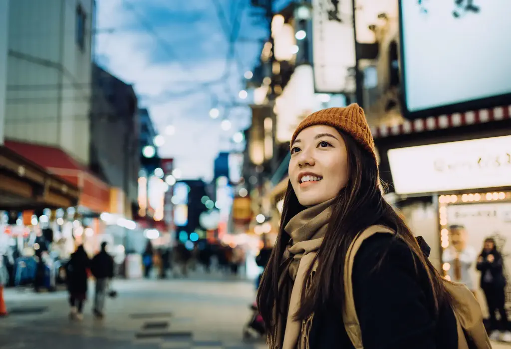 A smiling young Asian female traveller, sporting a beanie, scarf, and black coat, delightfully explores and strolls through a bustling local city street, soaking in the vibrant atmosphere of her surroundings.