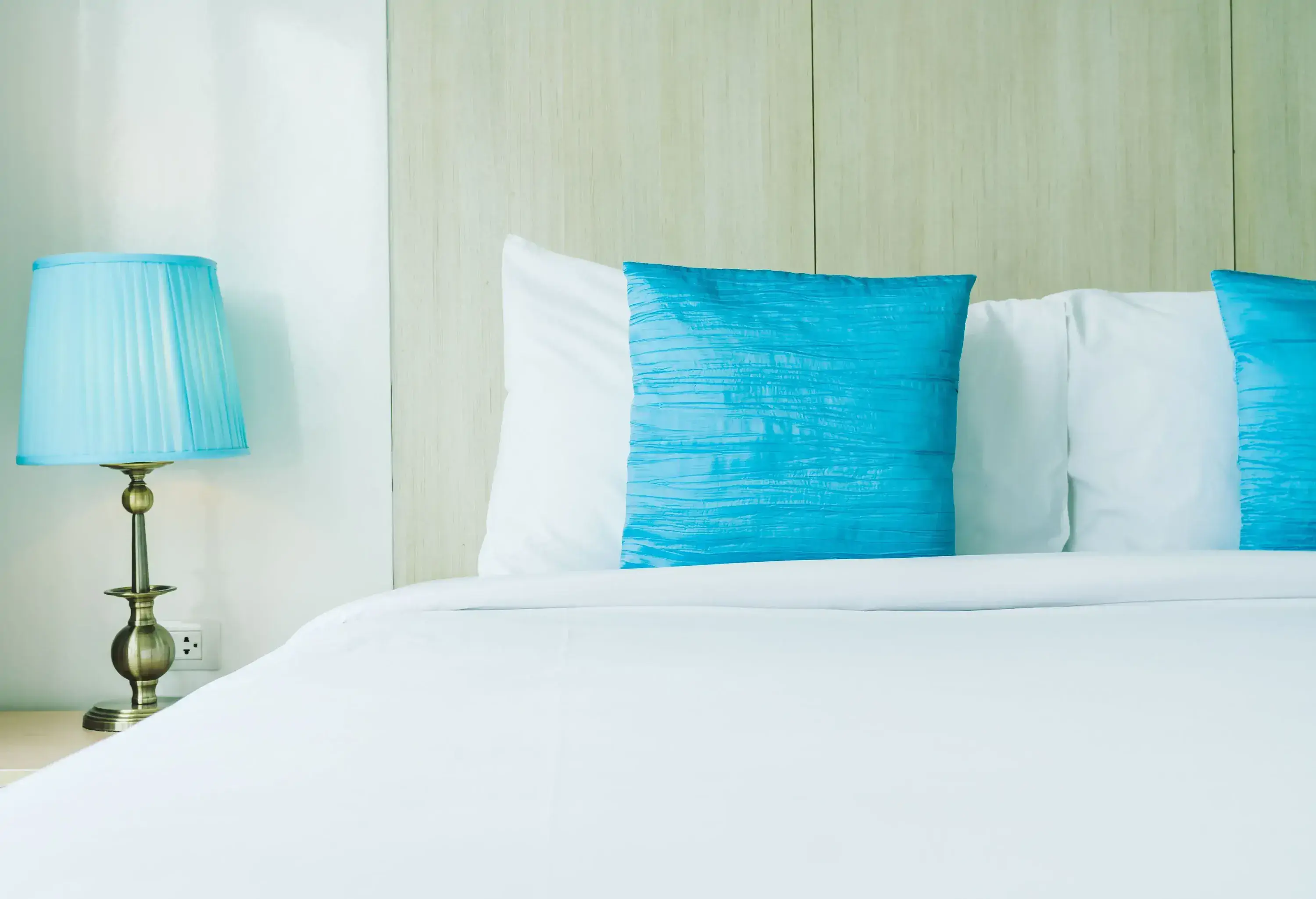 A bed with white beddings and light blue pillows beside a lavender lamp.