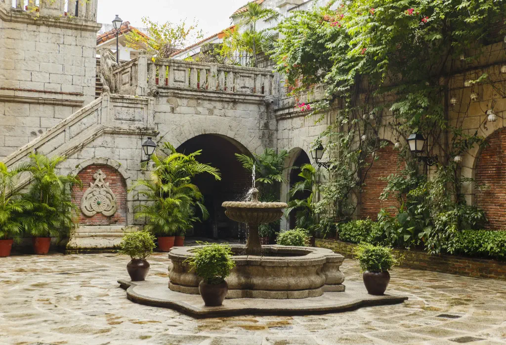 An ancient building's patio with potted plants surrounding the fountain and along the archway beneath the stairs.