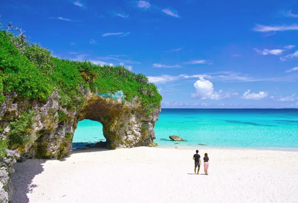 Two people walking across a sandy white beach with a natural arch.