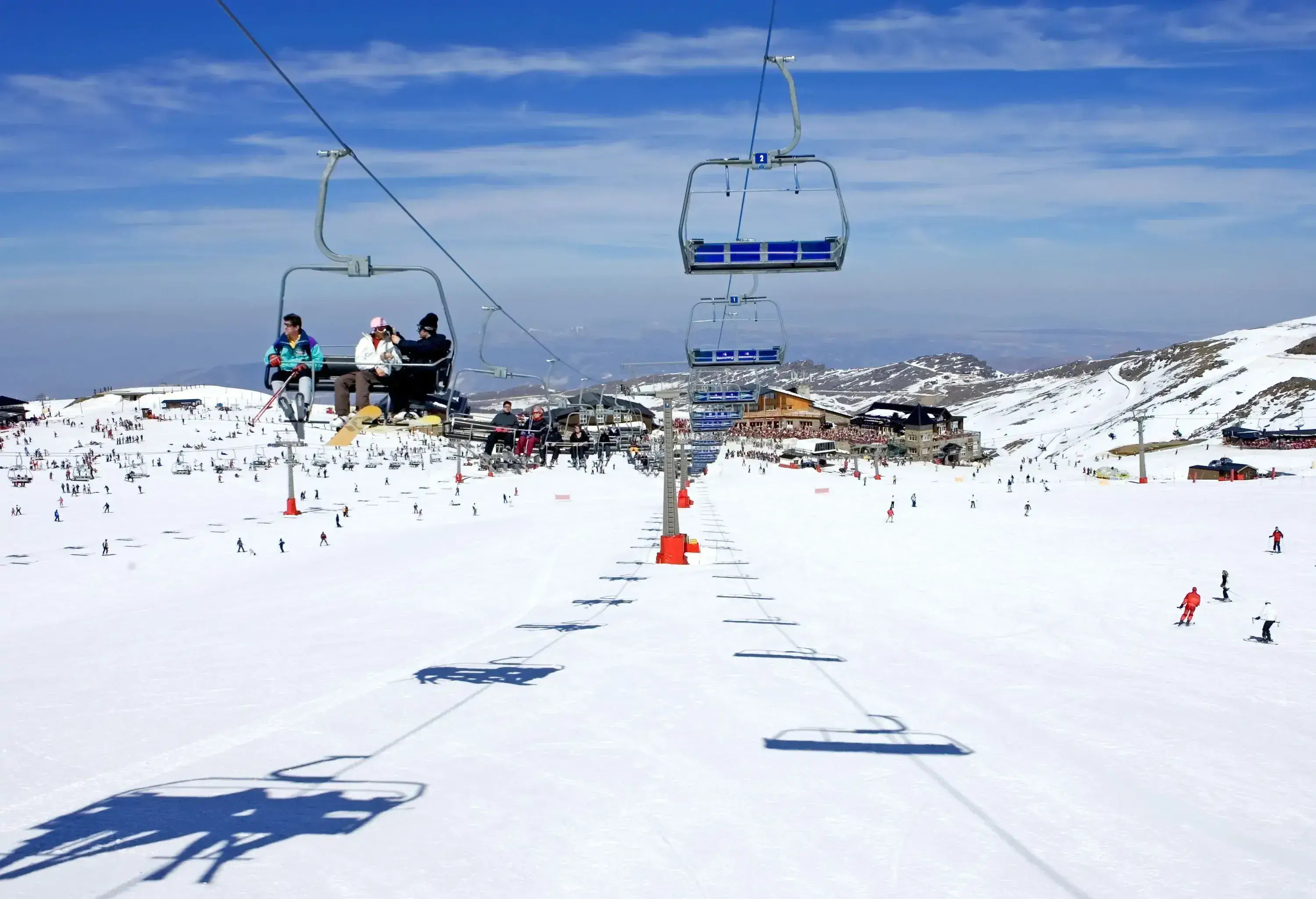 People sitting on a ski lift above people skiing across a vast snowy landscape in a rolling snowy valley.