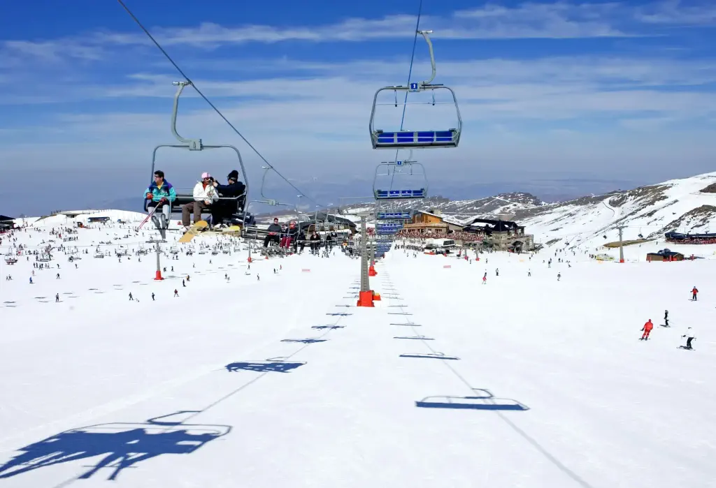 People sitting on a ski lift above people skiing across a vast snowy landscape in a rolling snowy valley.