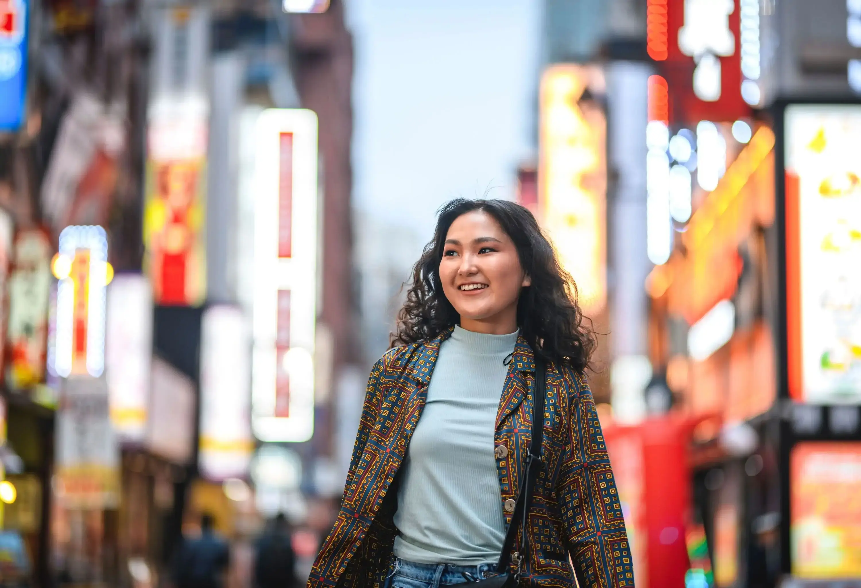 A smiling young woman walks through a city street bustling with bright lights.