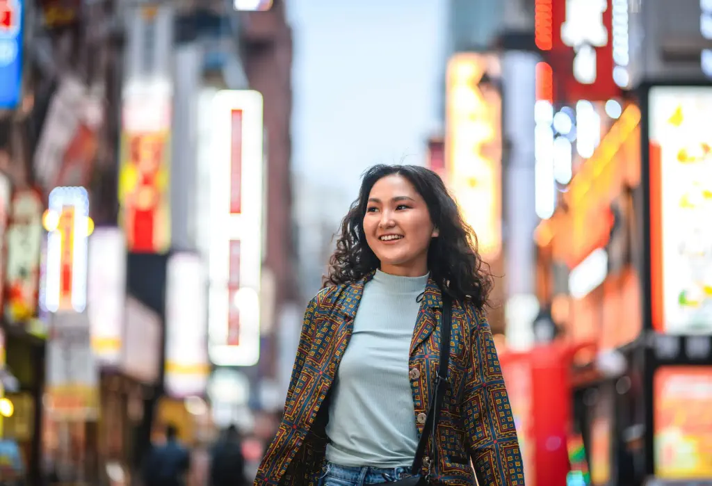 A smiling young woman walks through a city street bustling with bright lights.