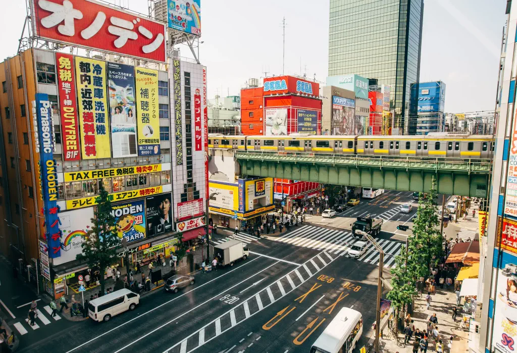 An avenue adorned with shops on both sides, featuring vibrant and colourful advertisements, while an elevated train gracefully glides along the tracks above.