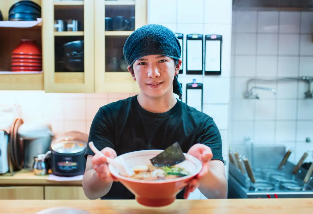 Japanese chef looking into the camera as he serves a bowl of freshly cooked ramen