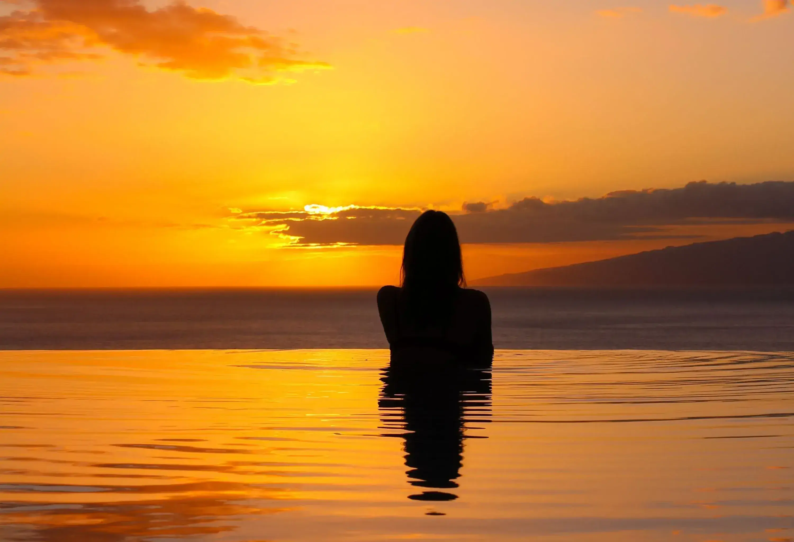 A woman bathing in an infinity pool under the orange sunset sky.