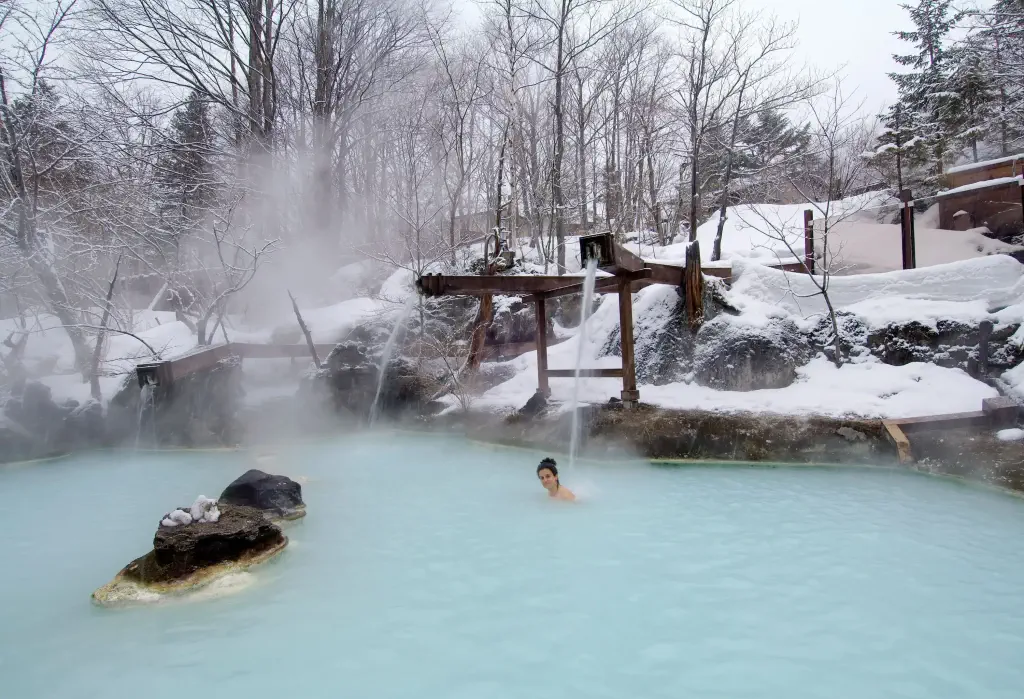 A woman dips into a steaming outdoor hot spring surrounded by frosted trees and snow-covered landscapes.
