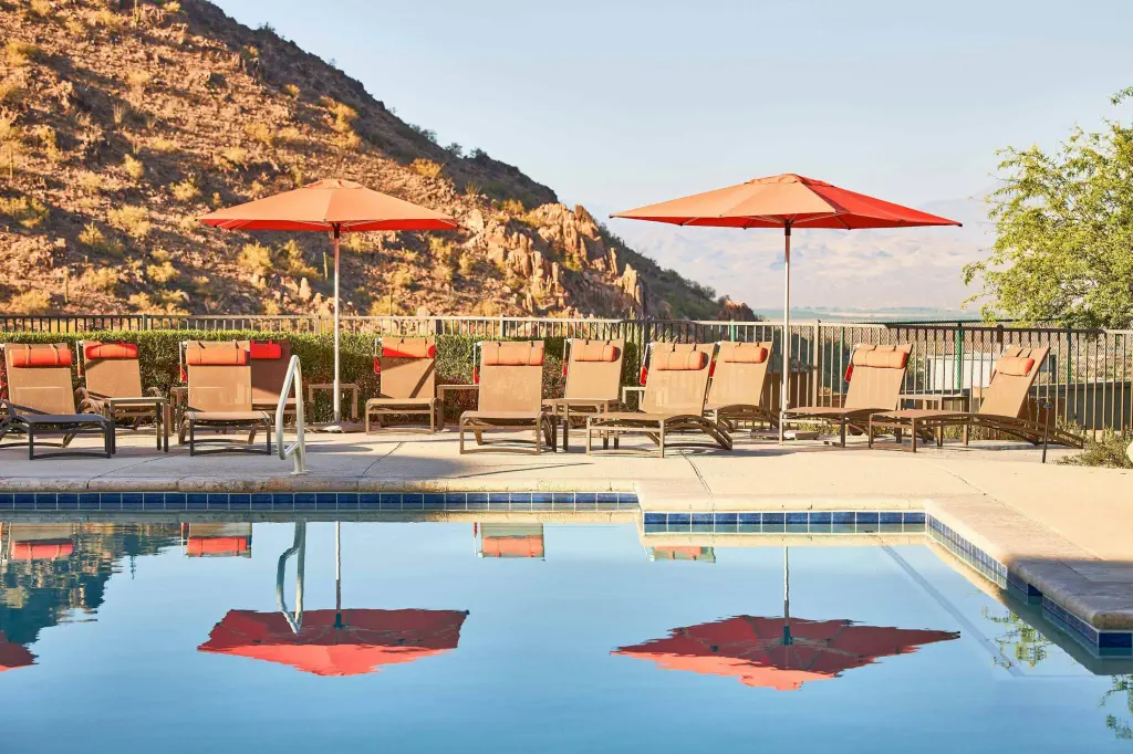 Poolside chairs and orange umbrellas reflected in a calm pool, set against a backdrop of sunlit rocky hills and a clear sky, evoke a tranquil, relaxing atmosphere.