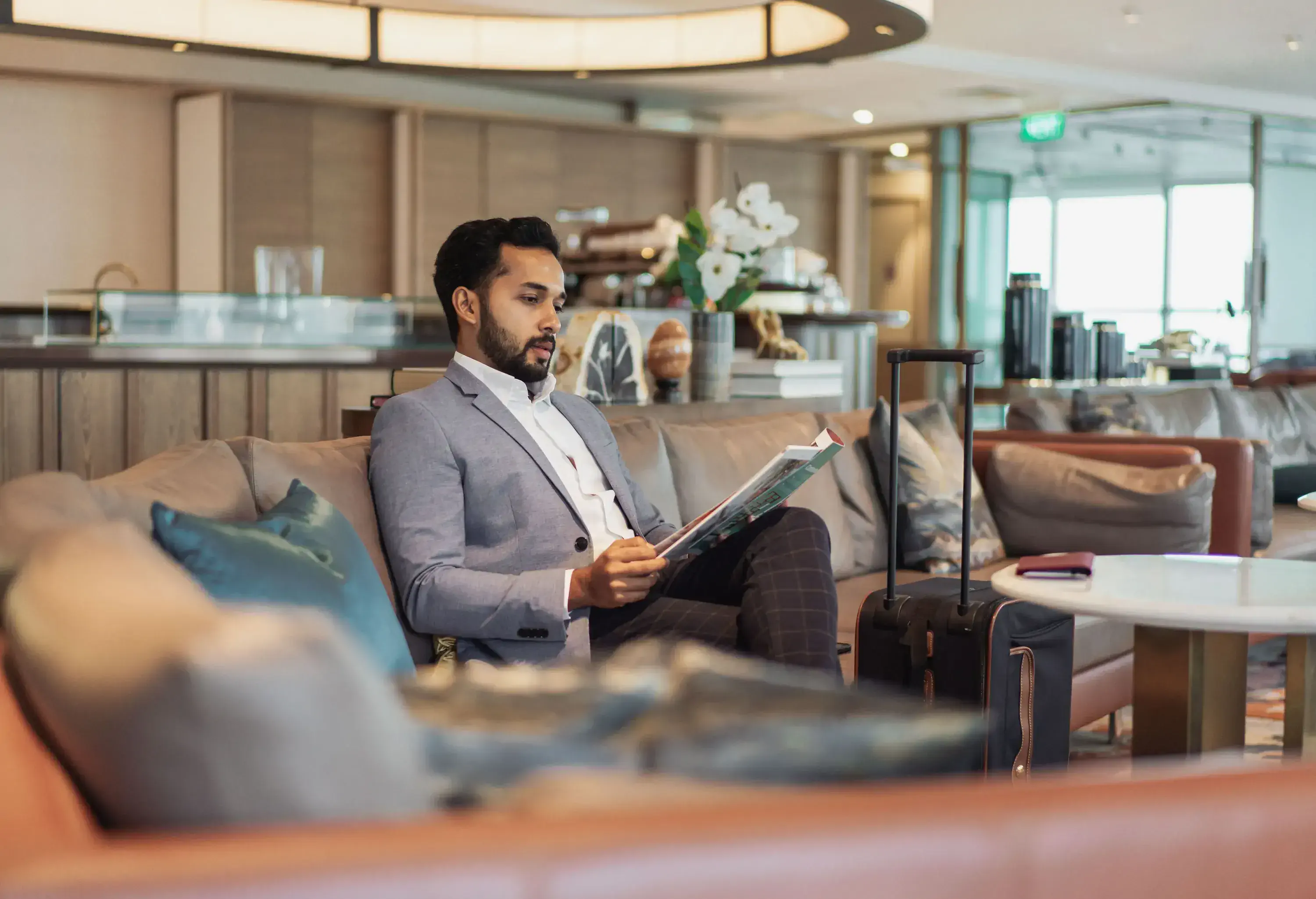 A man in a suit jacket sits on a sofa reading a magazine with a suitcase beside him in a modern lounge.