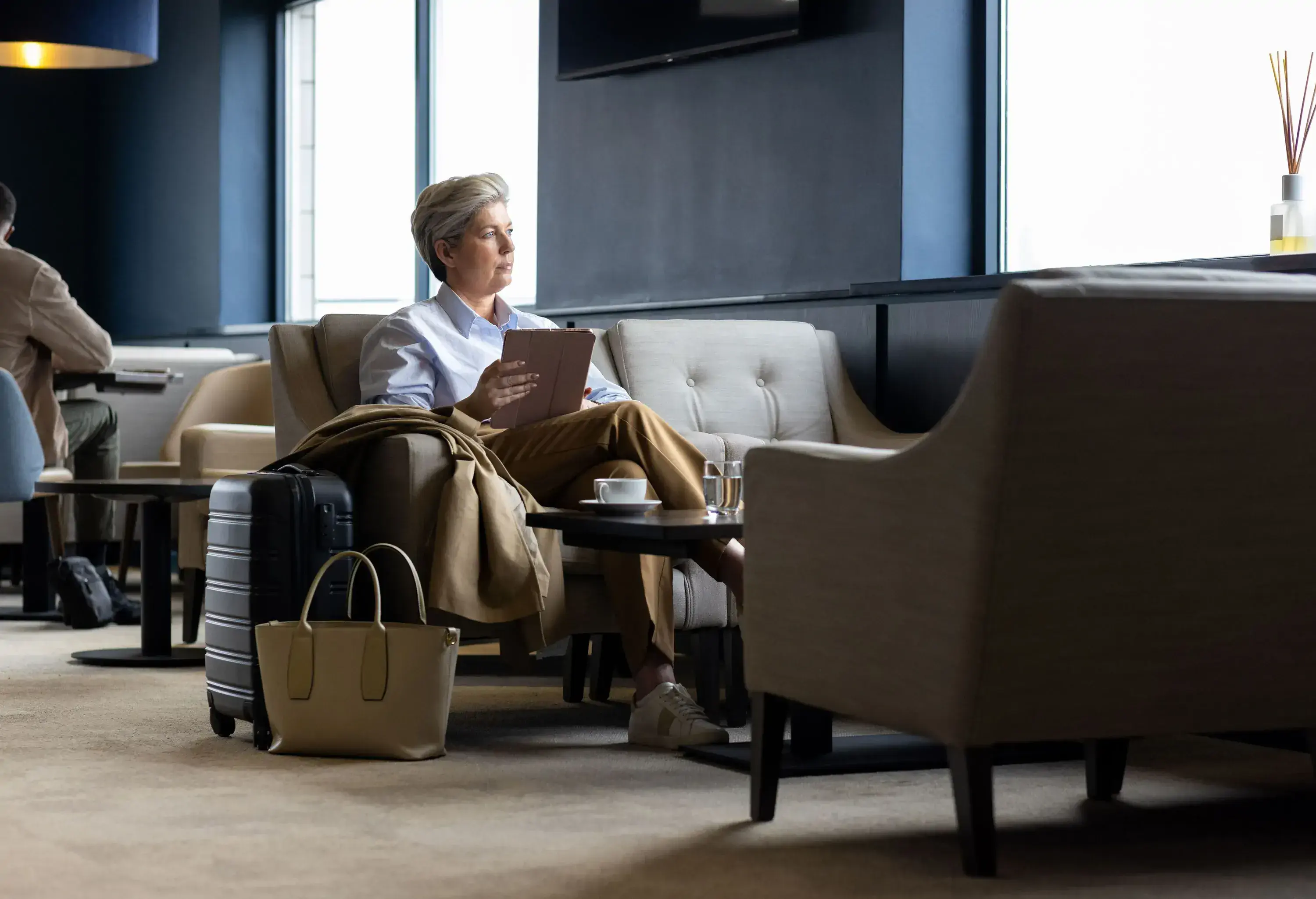 A full shot of a mature female adult sitting in an airport VIP lounge. She is holding a digital tablet sitting next to her carry-on luggage waiting for her gate to be called. The airport is located in the North East of England...Videos are available similar to this scenario.