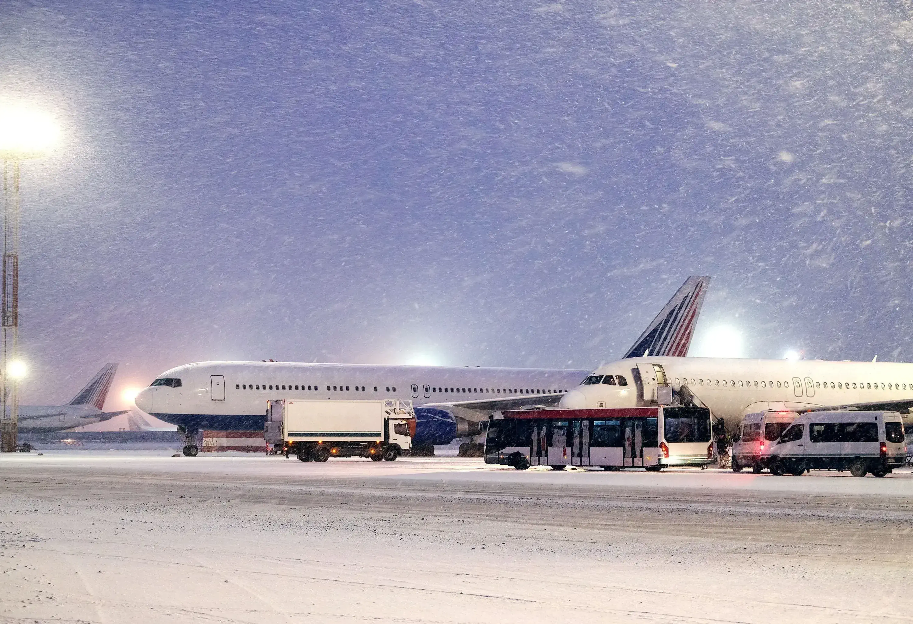 A snow-covered tarmac with two large airplanes and several smaller vehicles under strong overhead lighting in a heavy snowfall.
