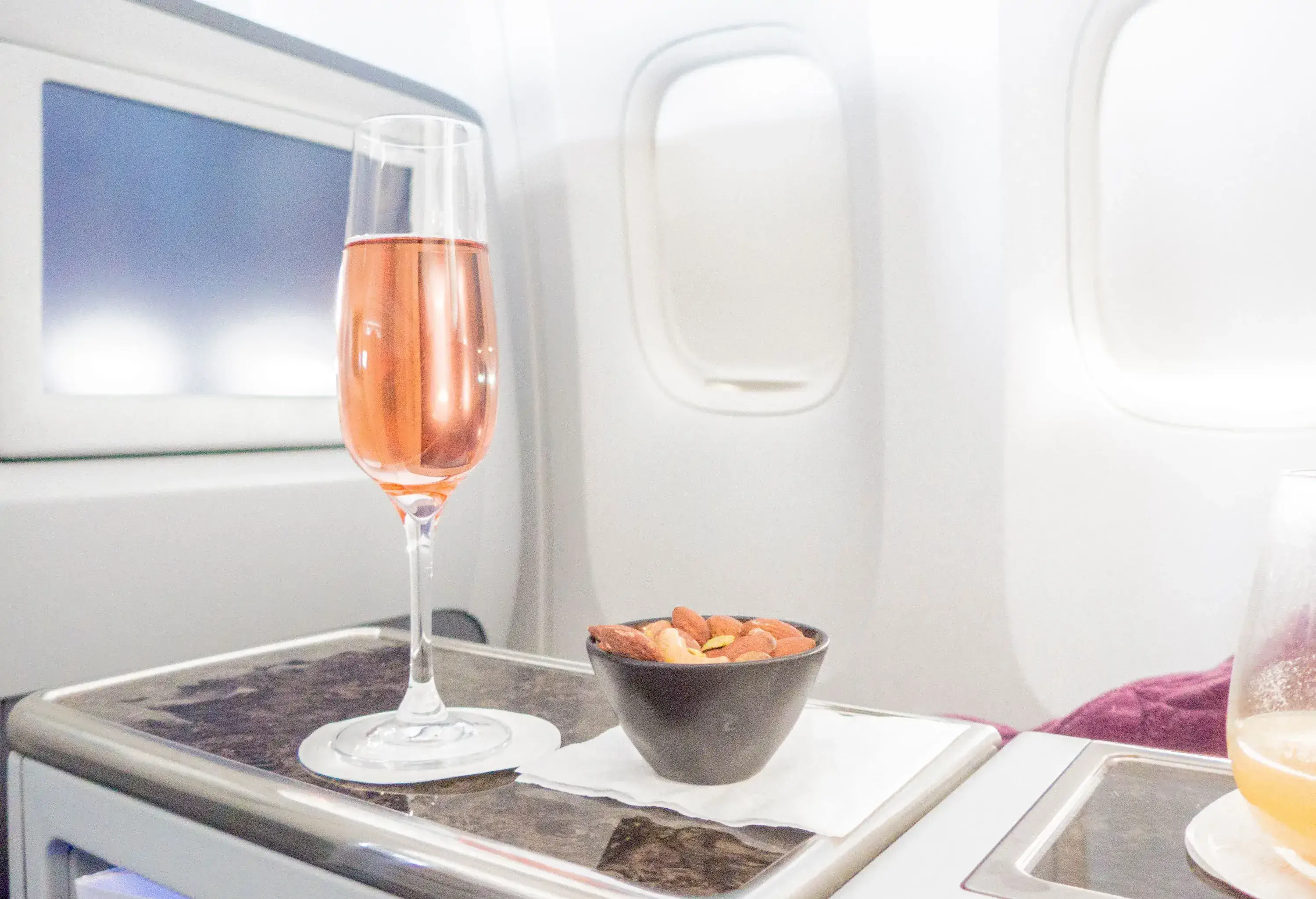 In the luxurious setting of a first-class cabin during a flight, an exquisite glass of pink champagne accompanies a selection of elegantly heated nuts, creating an indulgent and refined in-flight experience.