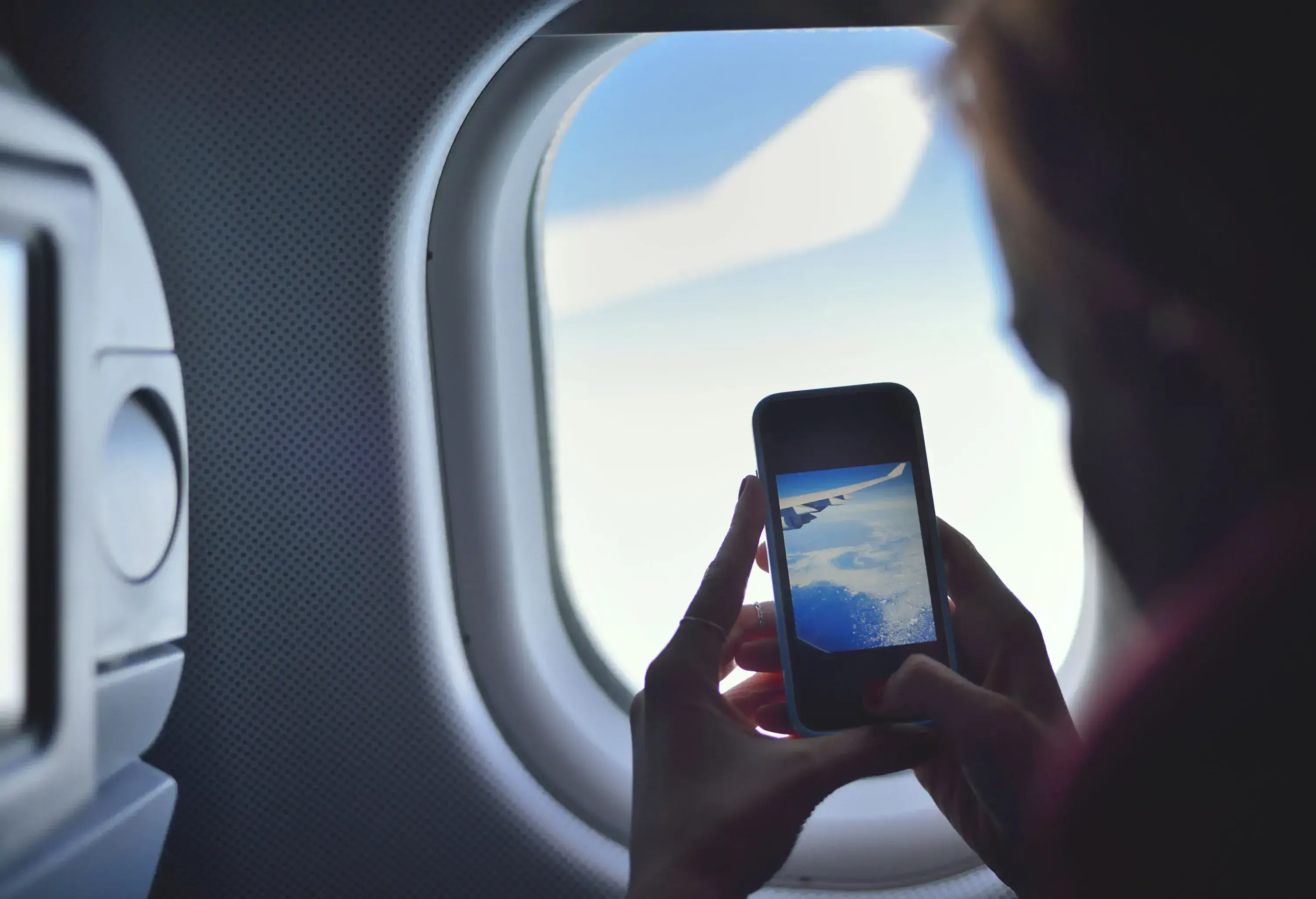 A passenger seated inside an aircraft by the window captures a photo of the white clouds using her smartphone.