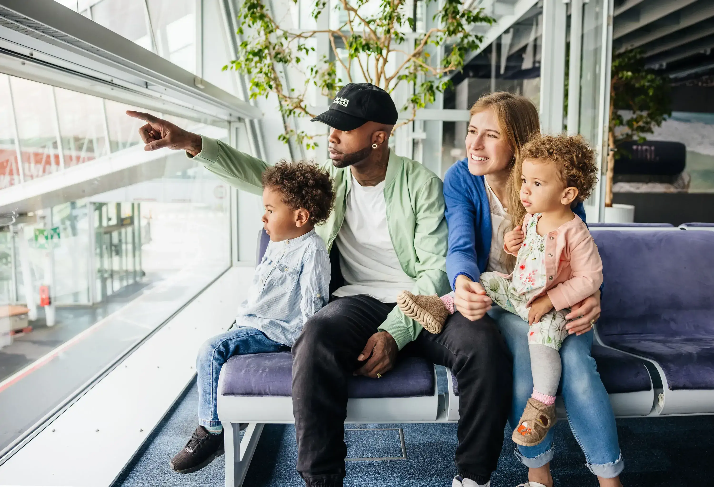 A family of four seated on an airport bench, looking intently out the large window.