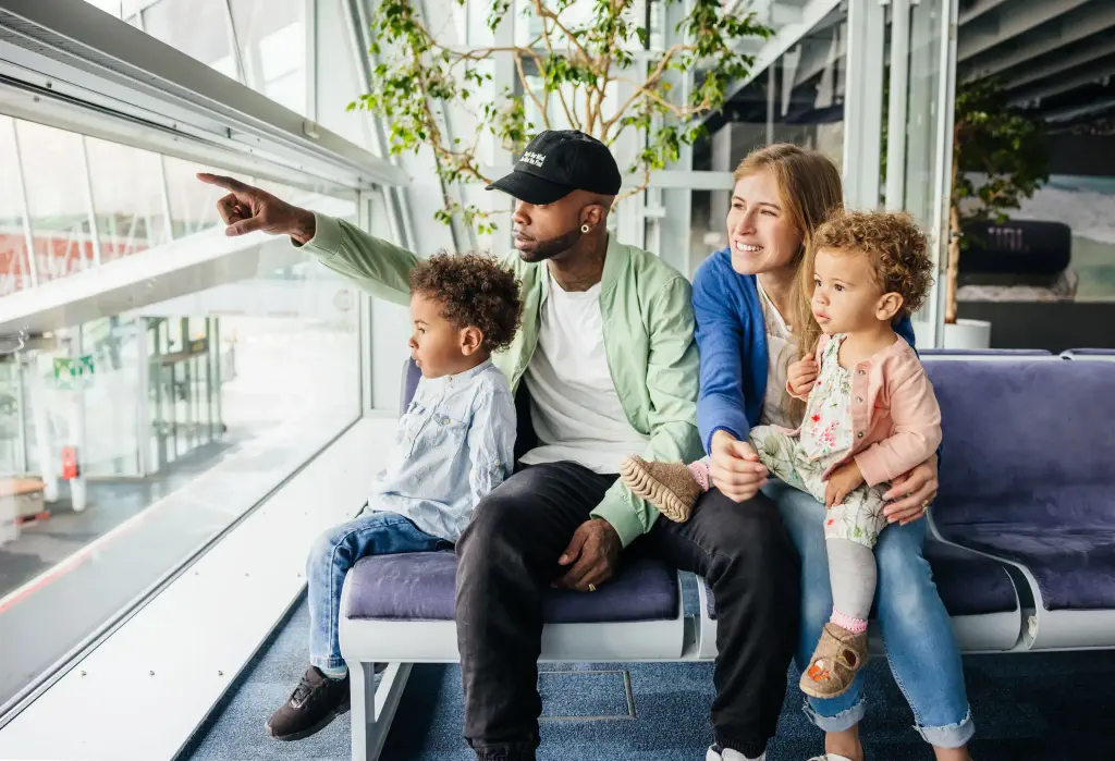 A family of four seated on an airport bench, looking intently out the large window.