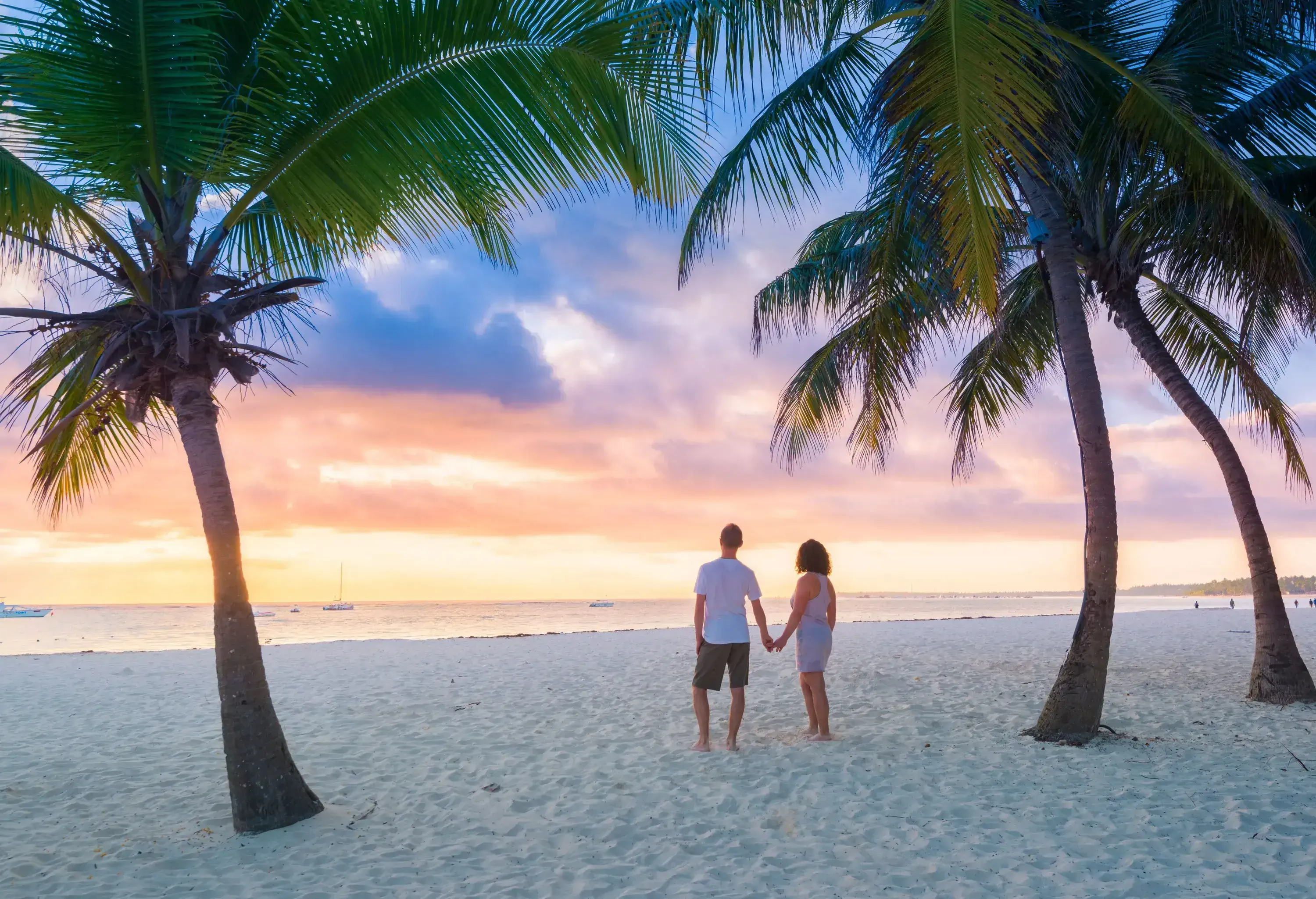A couple holding hands as they stand under the palm trees while admiring the sunset on the beach.