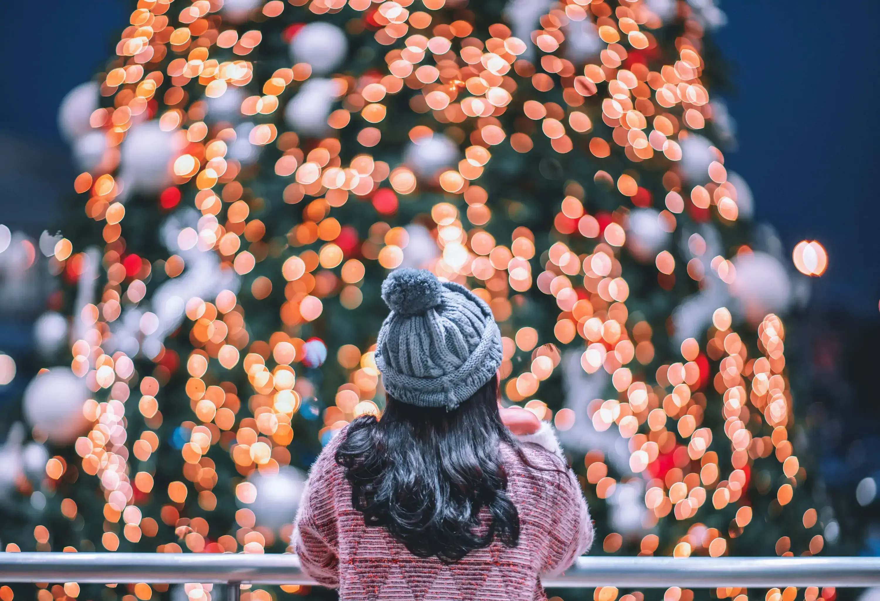 Woman with long hair and a knitted hat leaning on a railing to gaze at a Christmas tree.