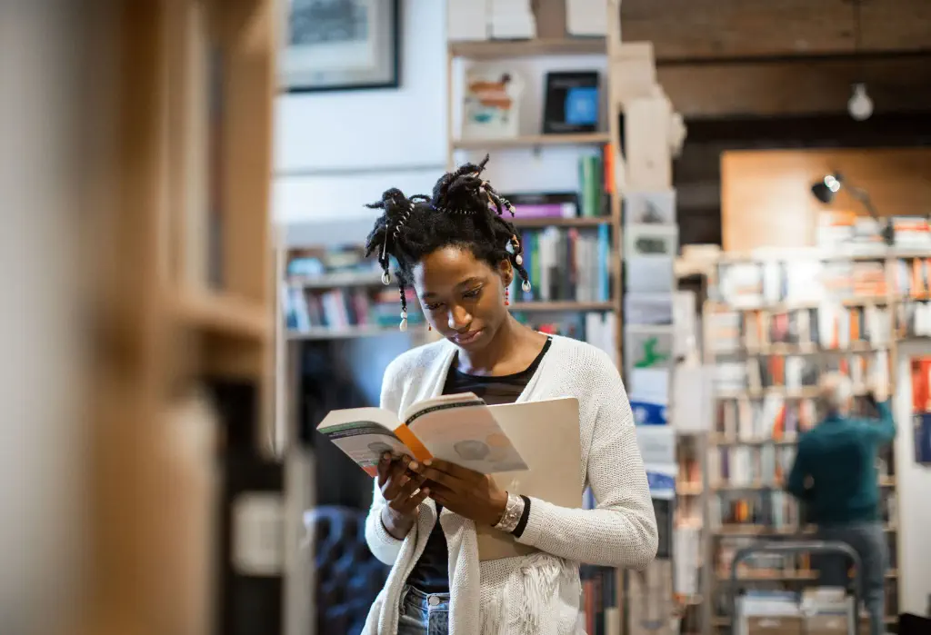 A young woman is engrossed in reading a book while standing amidst the stacked shelves of a bustling bookstore.