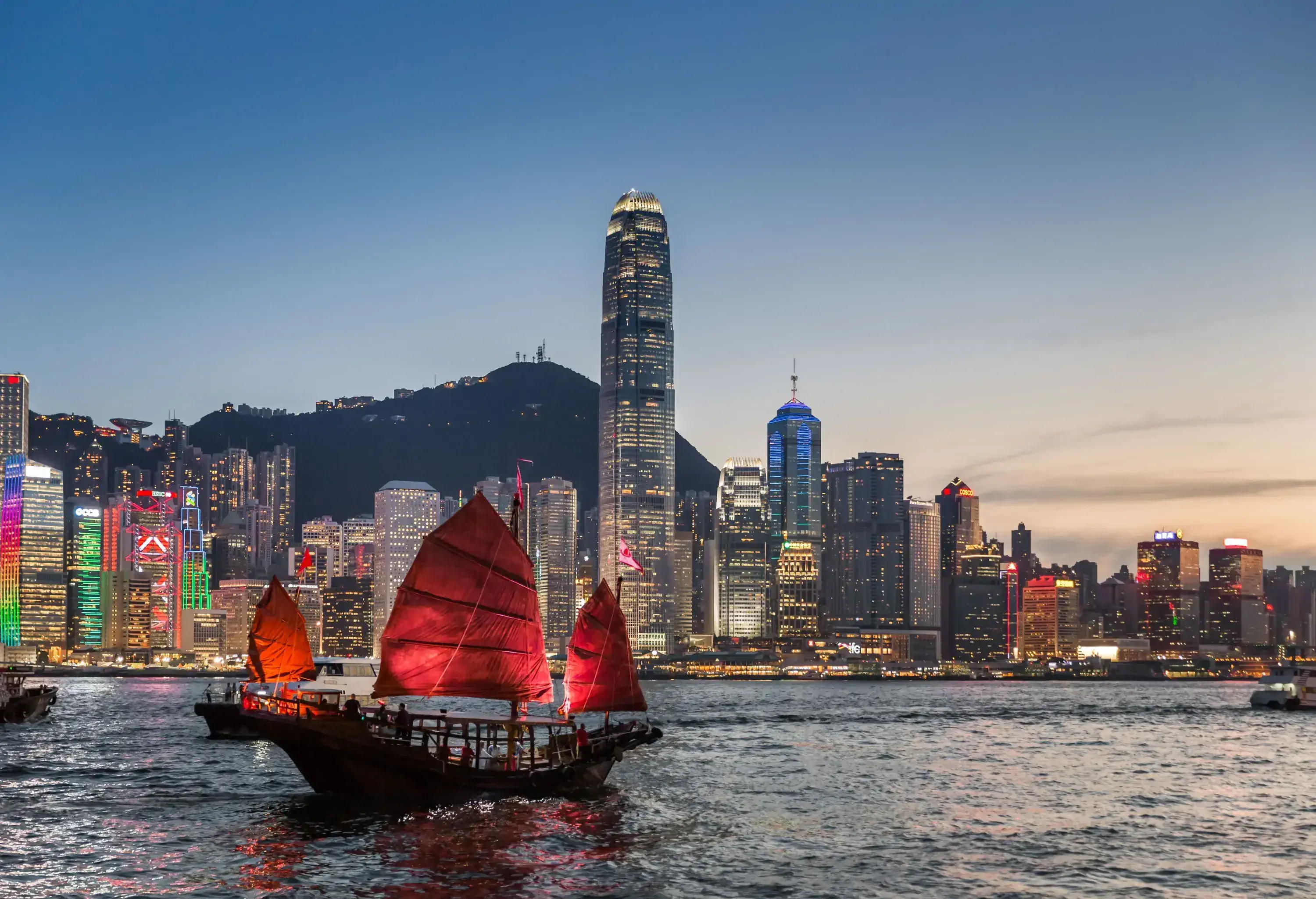 A traditional junk boat sails across the brightly lit modern city with towering buildings at twilight.