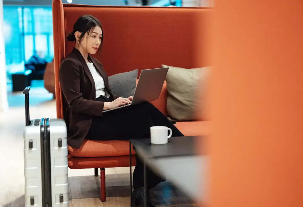 A young stylish woman uses a laptop while seated comfortably in an orange upholstered chair, with a white suitcase next to her.