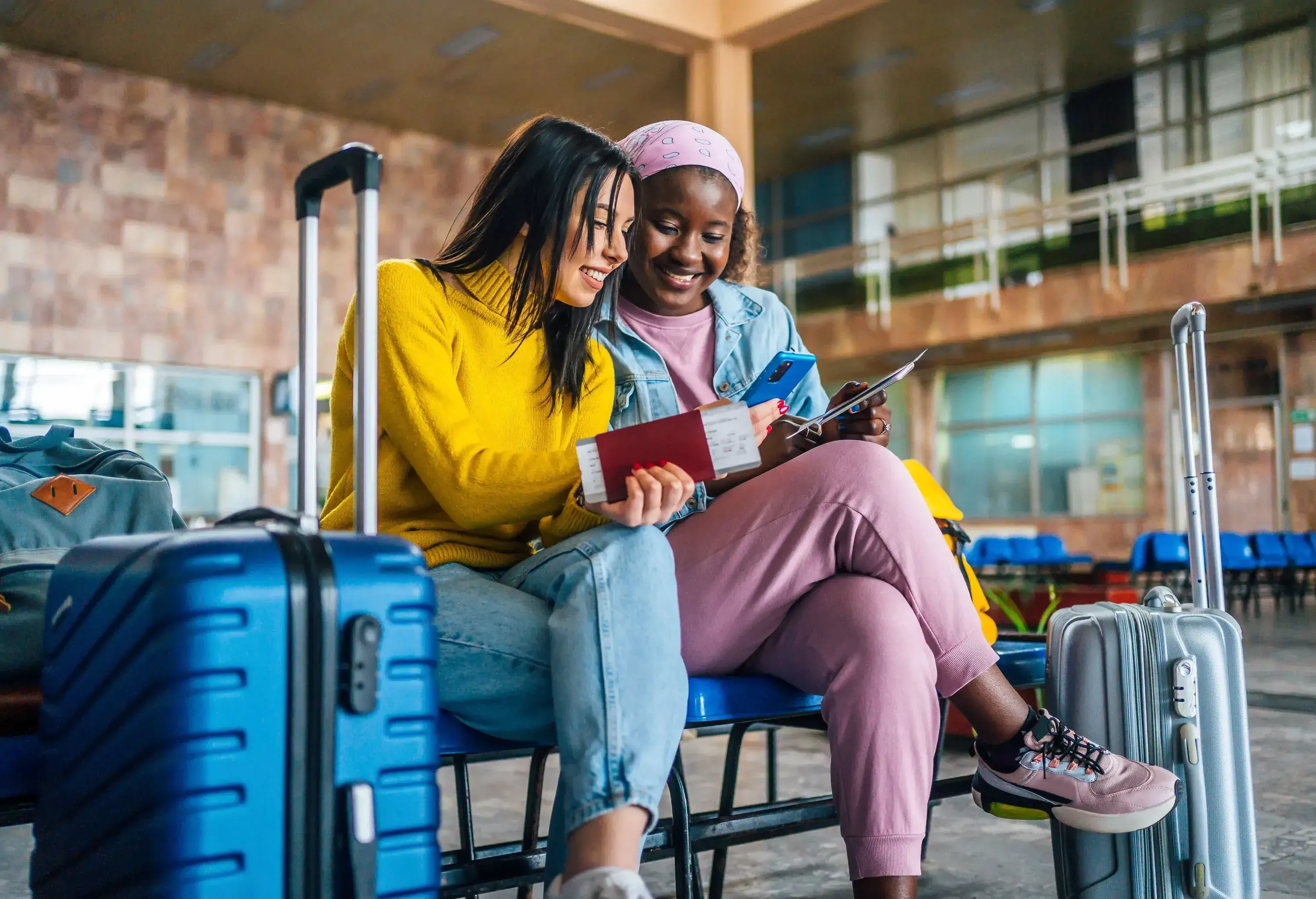 Two young women sitting in a waiting area with their luggage, looking at a phone and holding passports.
