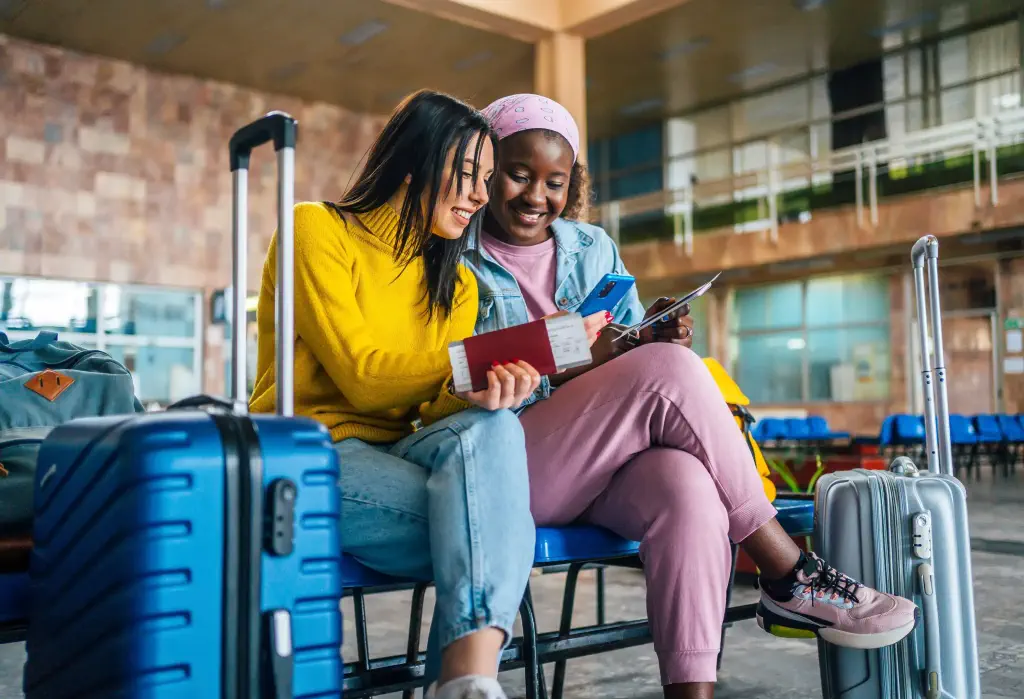 Two young women sitting in a waiting area with their luggage, looking at a phone and holding passports.