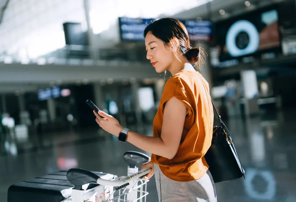 Young Asian woman using smartphone and pushing a luggage trolley with suitcases at airport terminal. Checking-in with mobile phone. Ready for a trip. Business travel. Travel and vacation concept
