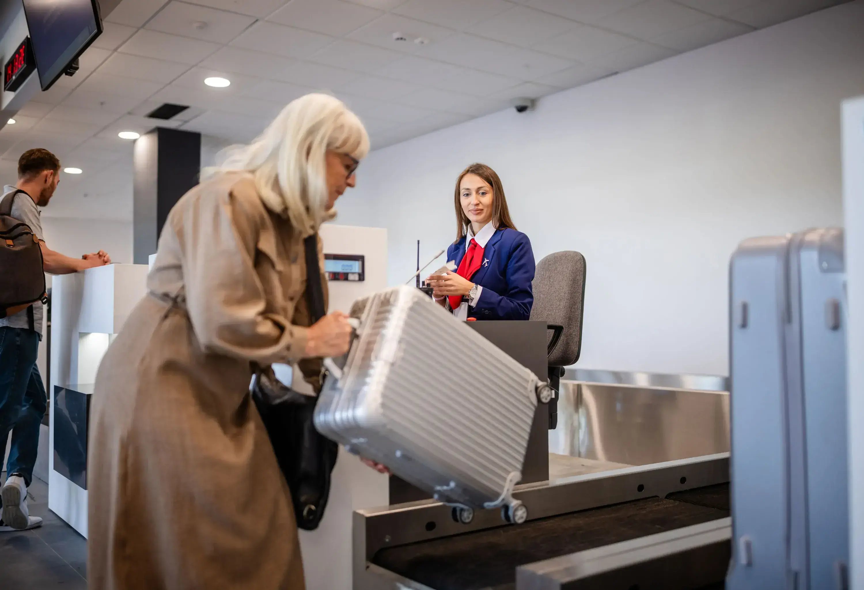 An elderly passenger carefully positions her suitcase onto the conveyor belt assisted by an airport staff member, illustrating baggage handling procedures at airports.