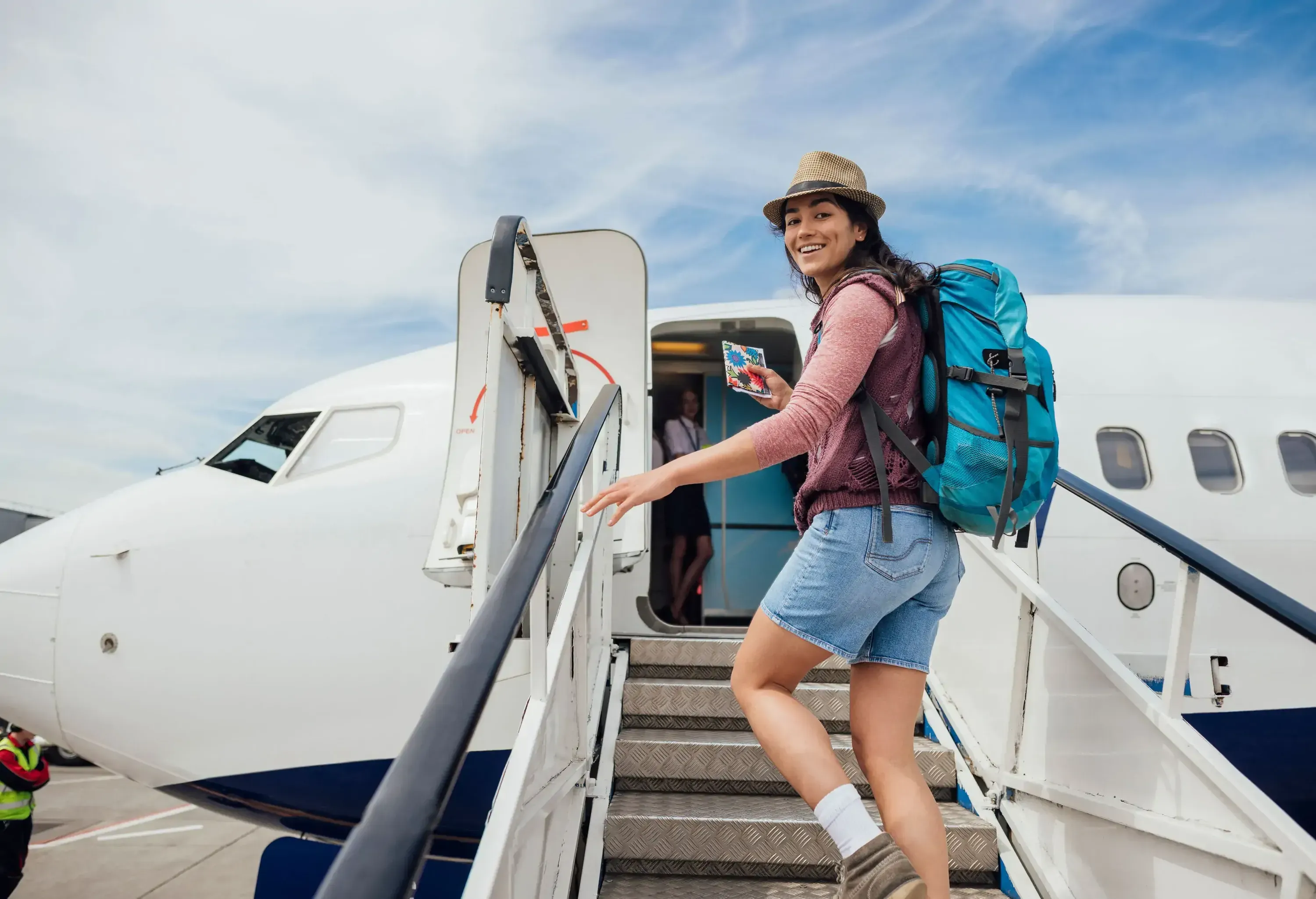 A young, smiling woman with a backpack and a hat climbing the stairs to board an airplane.