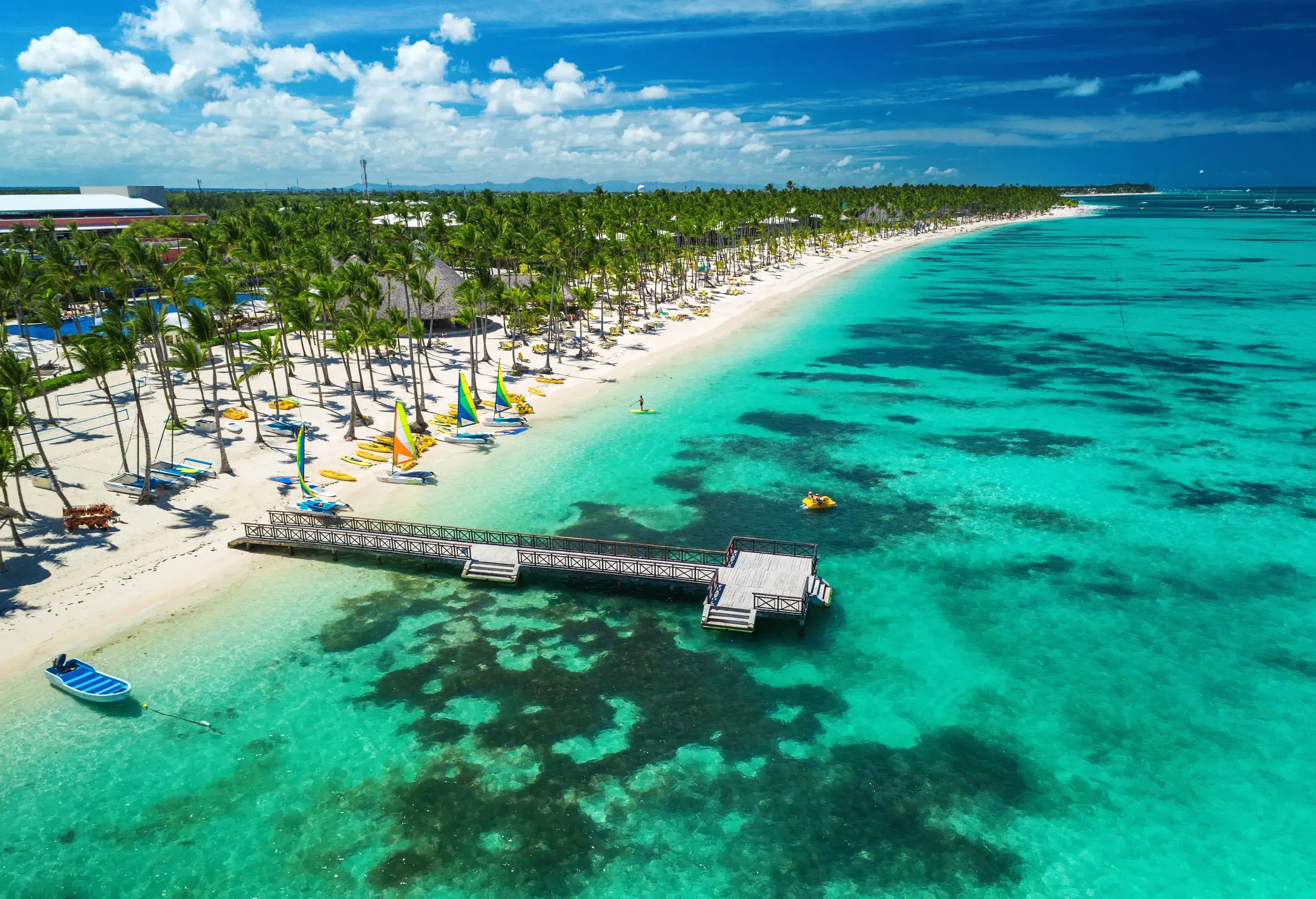 A jetty extending to the sea's crystalline shallow water in a white sandy beach full of lush palm trees.
