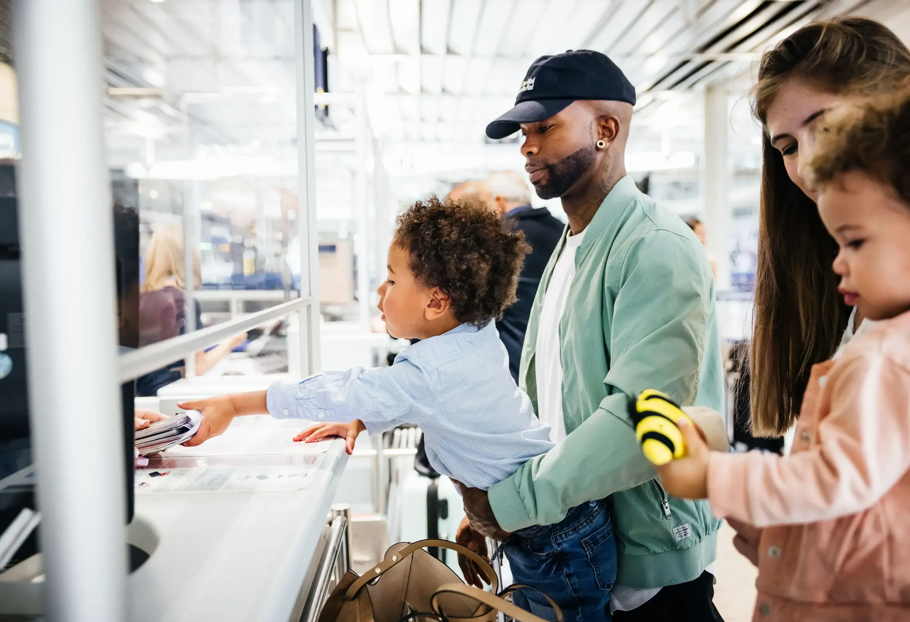 A family at an airport service point, with a man in a cap holding a small child who is reaching towards a counter.