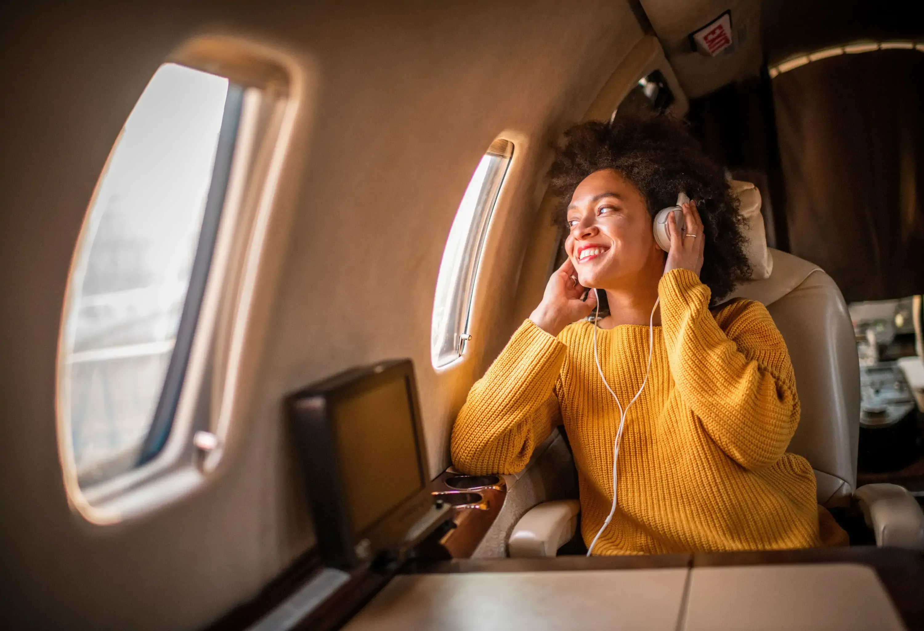 A smiling woman wearing headphones seated comfortably looks out the window of an aircraft.