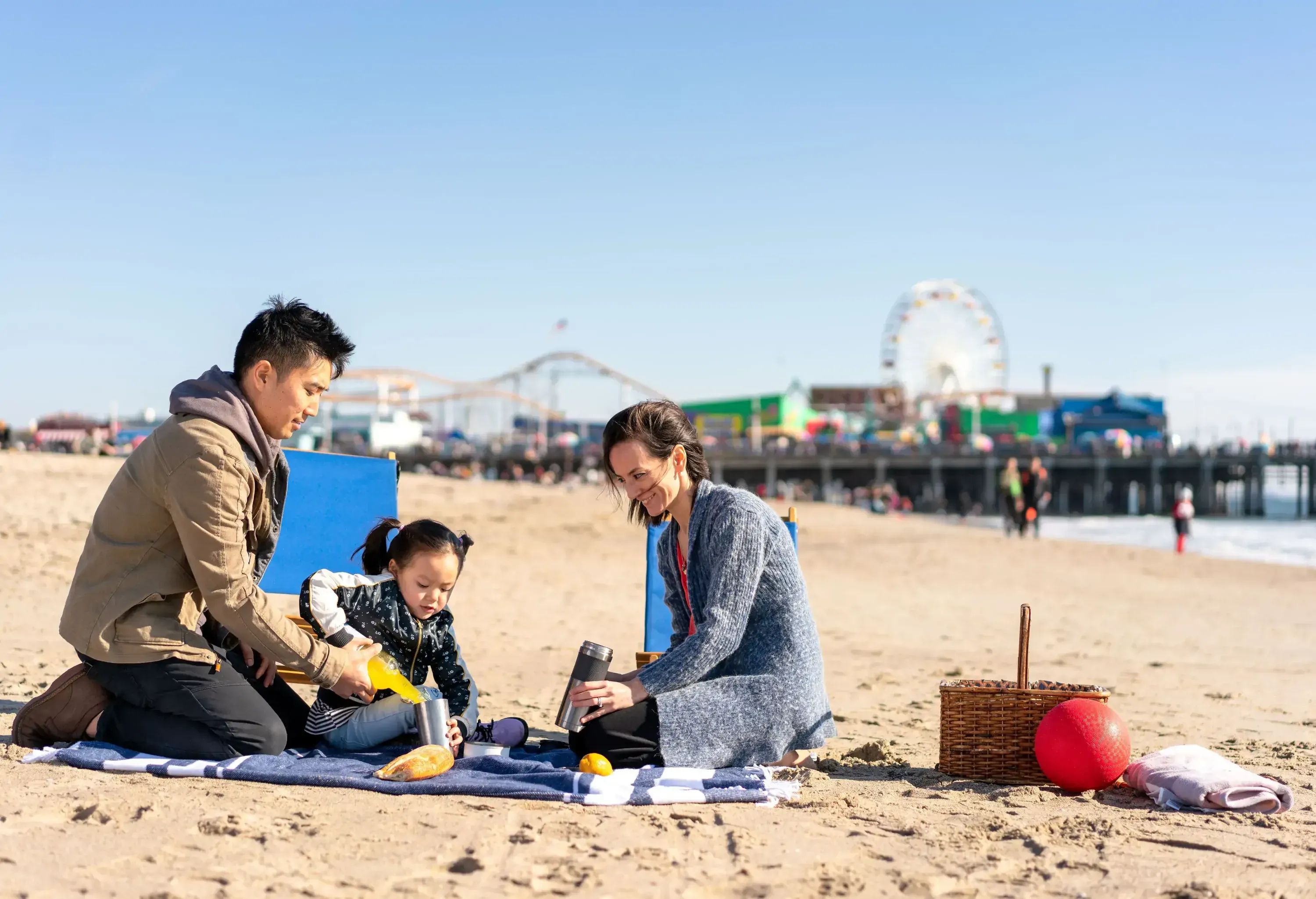 Three people sitting on a picnic rug on the beach, with the man assisting a kid in pouring a drink into a container while the woman observes.