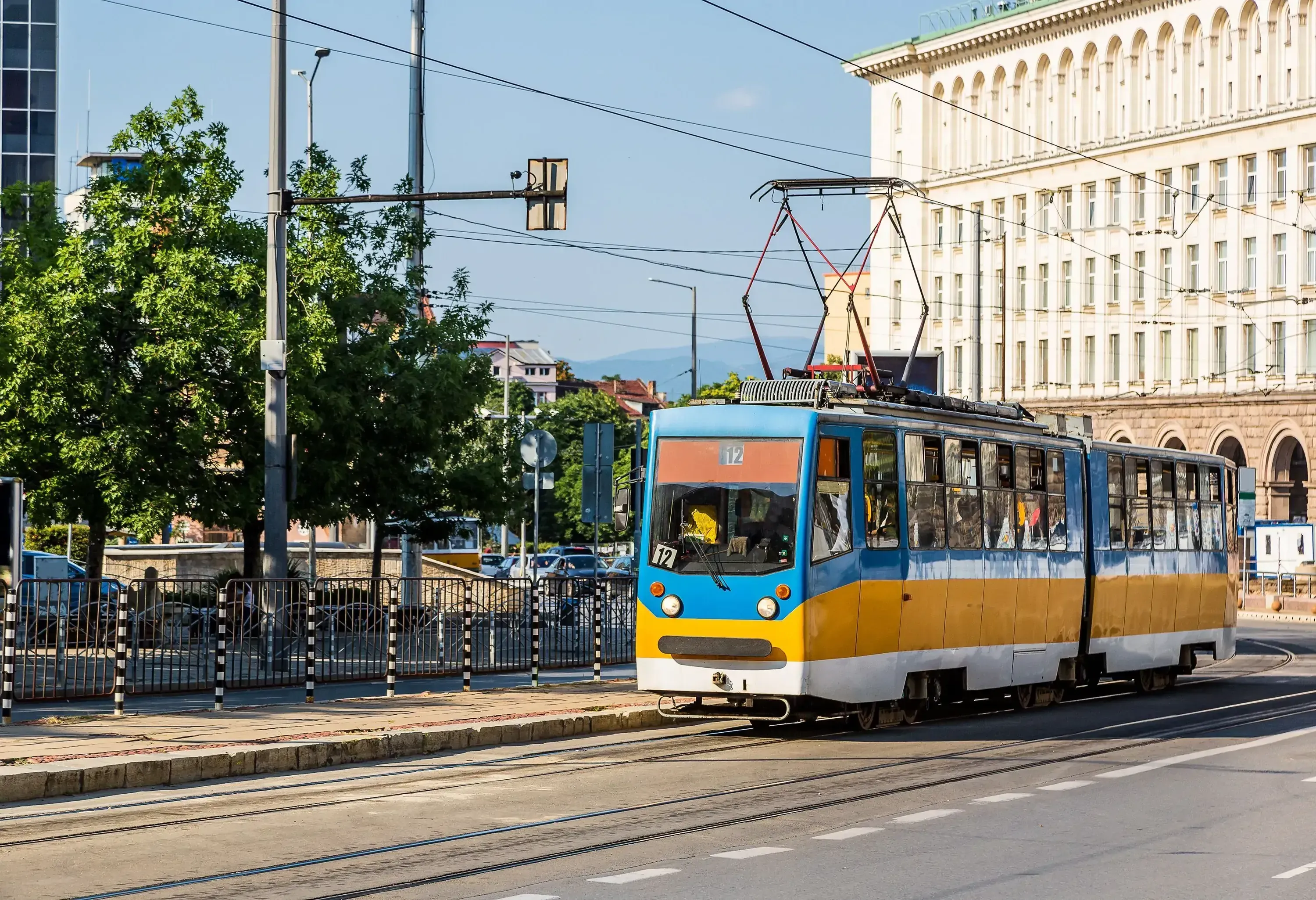 A vibrant yellow and blue tram gracefully glides along the street, with a charming park and buildings creating a picturesque backdrop.