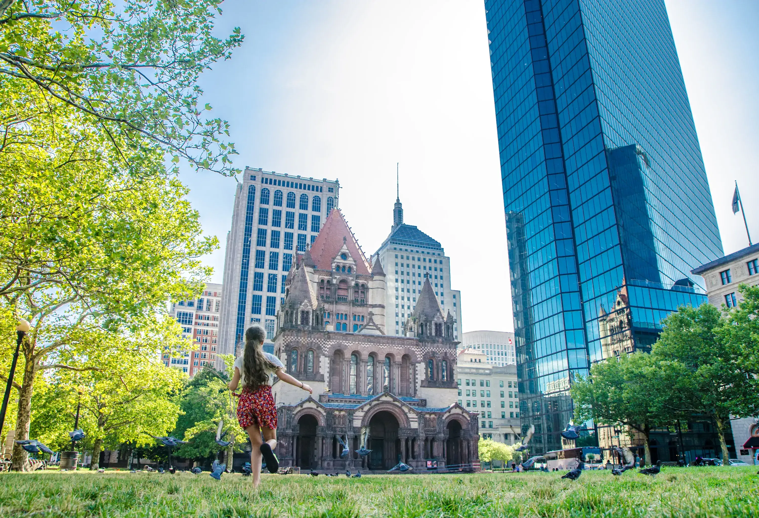 Boston Trinity Church with skyscrapers during summer day, with a teenager girl running after pigeons