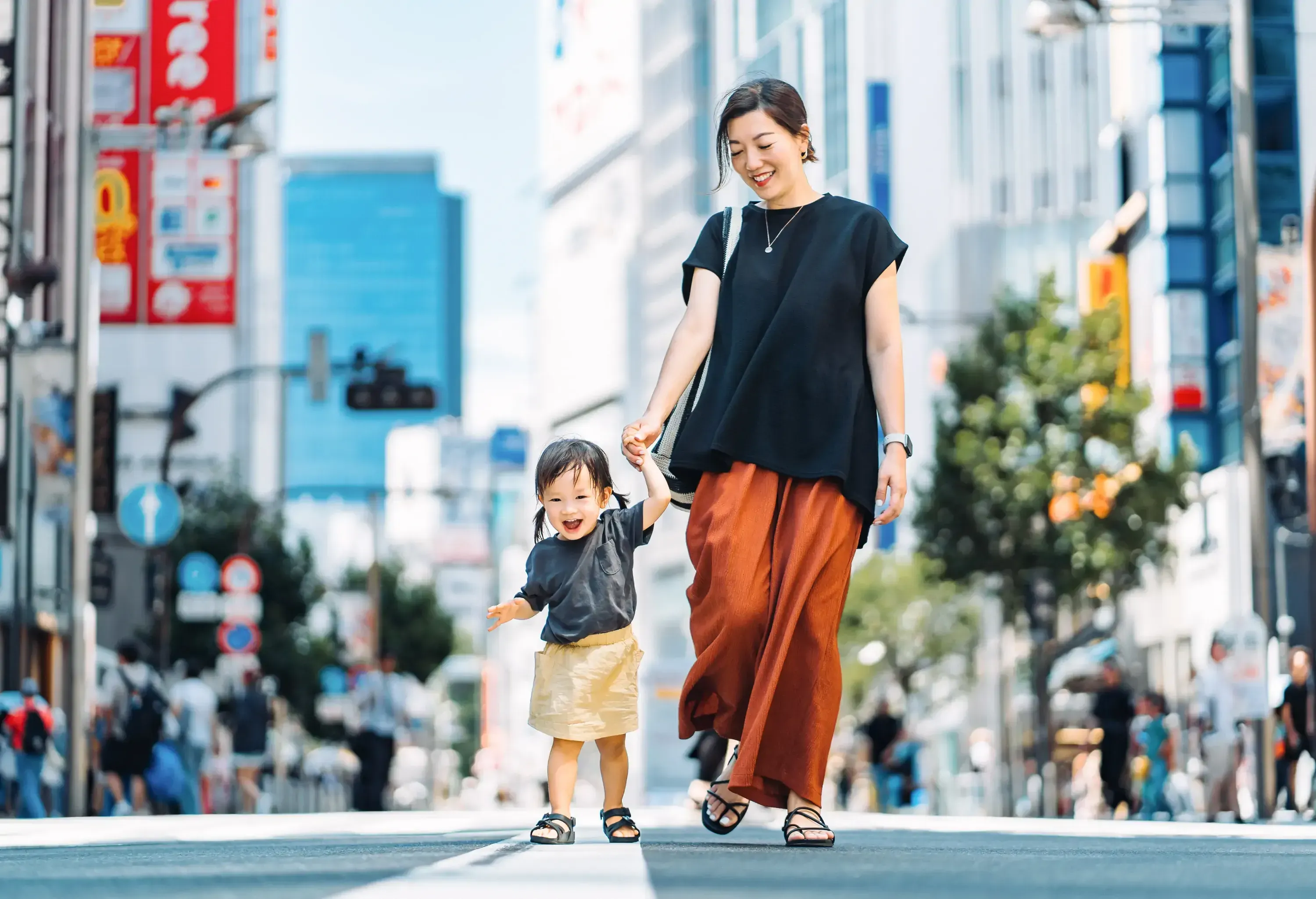 Joyful young Asian mother holding hands of her little daughter, walking in urban setting.