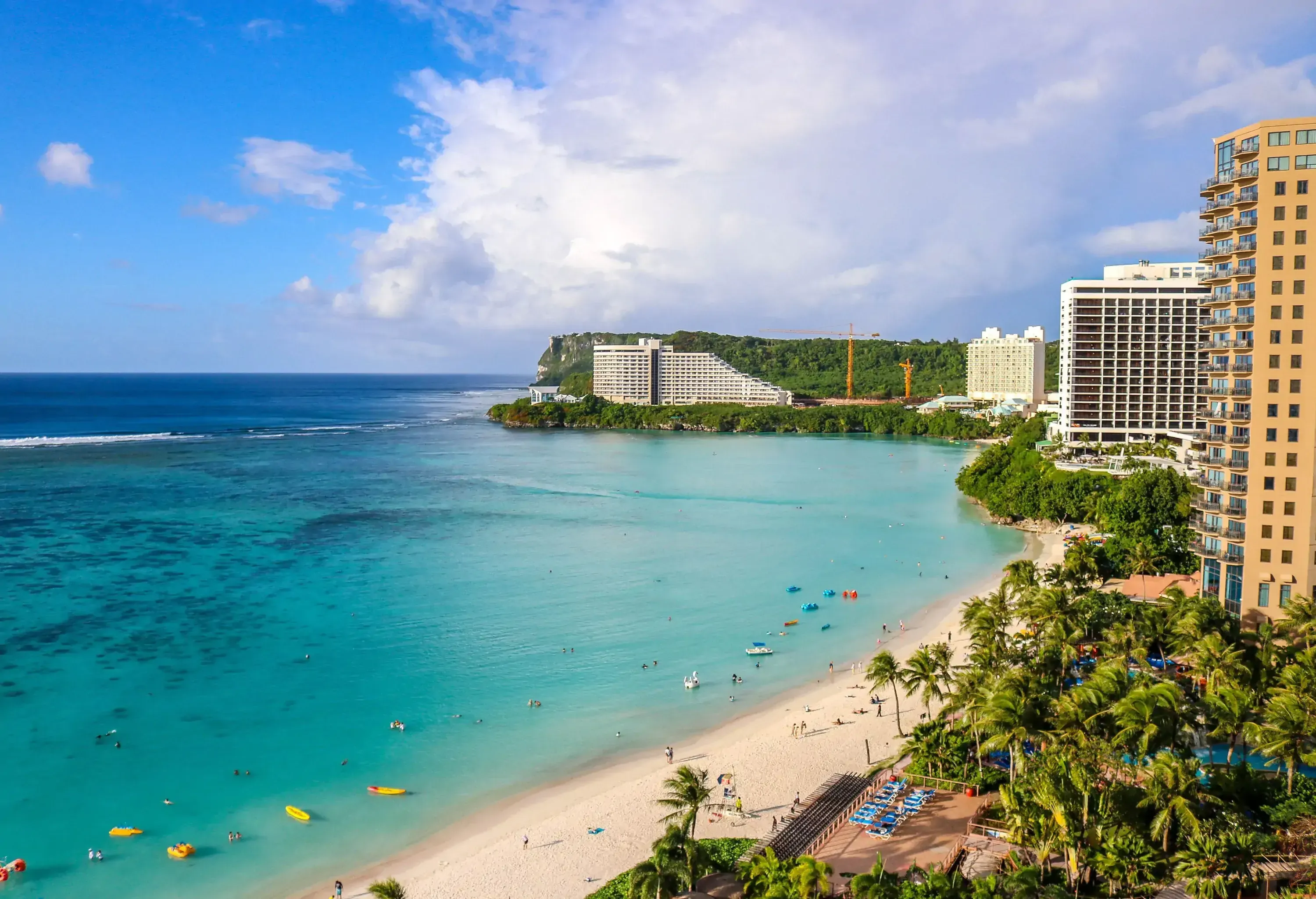 An uncrowded and relaxing turquoise beach with soft sand lined with lush plants and palm trees alongside the tall buildings.