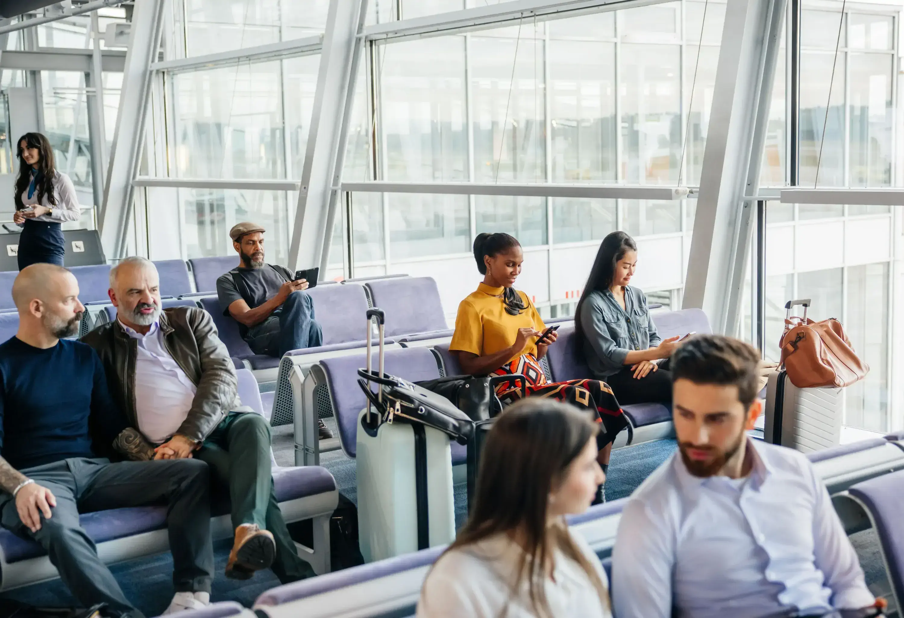Some passengers wait patiently in an airport lounge for their flights, sitting in chairs and passing the time chatting and looking at their smartphones.