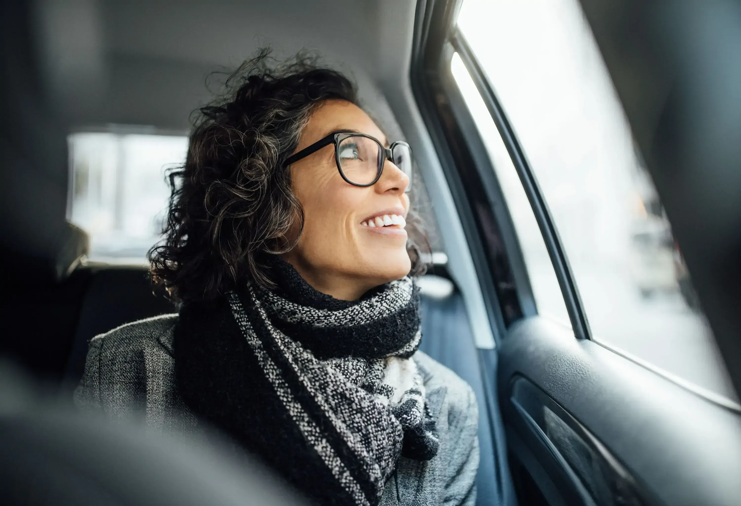 A smiling woman with glasses and a scarf looks out a car window.
